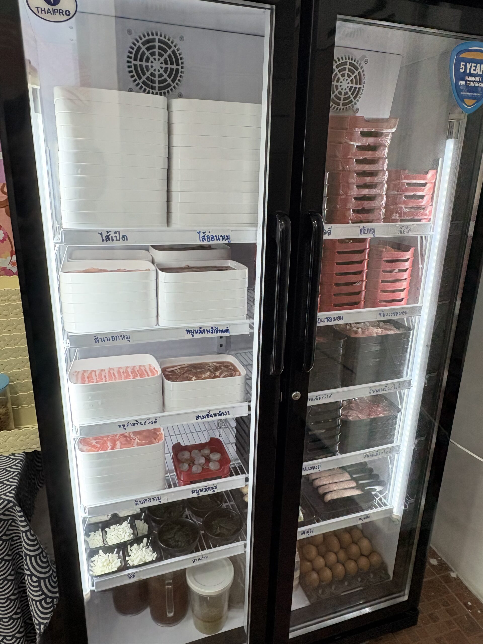ตู้แช่เย็นกระจกใสแบรนด์ THAIPRO จัดเรียงวัตถุดิบชาบูหมูหลากชนิด เช่น ไส้เป็ด ไส้อ่อนหมู สันนอกหมู หมูหมักพริกไทยดำ สามชั้นหมักรา ลูกชิ้น ไข่ไก่ และผักต่างๆ ในถาดขาวและแดง