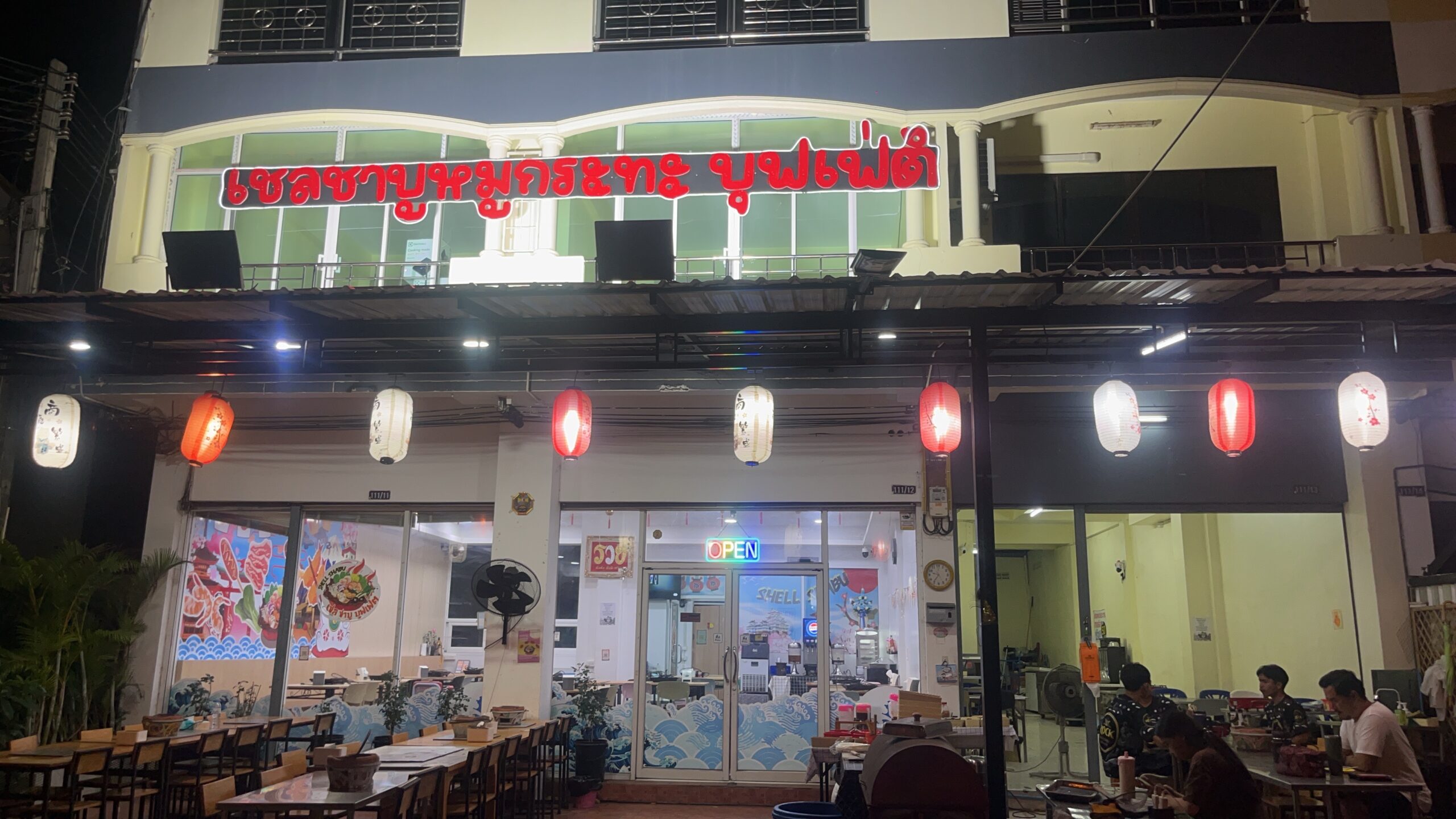 ร้านเชลชาบูหมูกระทะบุฟเฟ่ต์ ถ่ายตอนกลางคืน มีโคมไฟญี่ปุ่นสีแดงและสีขาวประดับหน้าร้าน ป้ายไฟสีแดงชื่อร้าน และป้ายไฟ OPEN เปิดให้บริการ