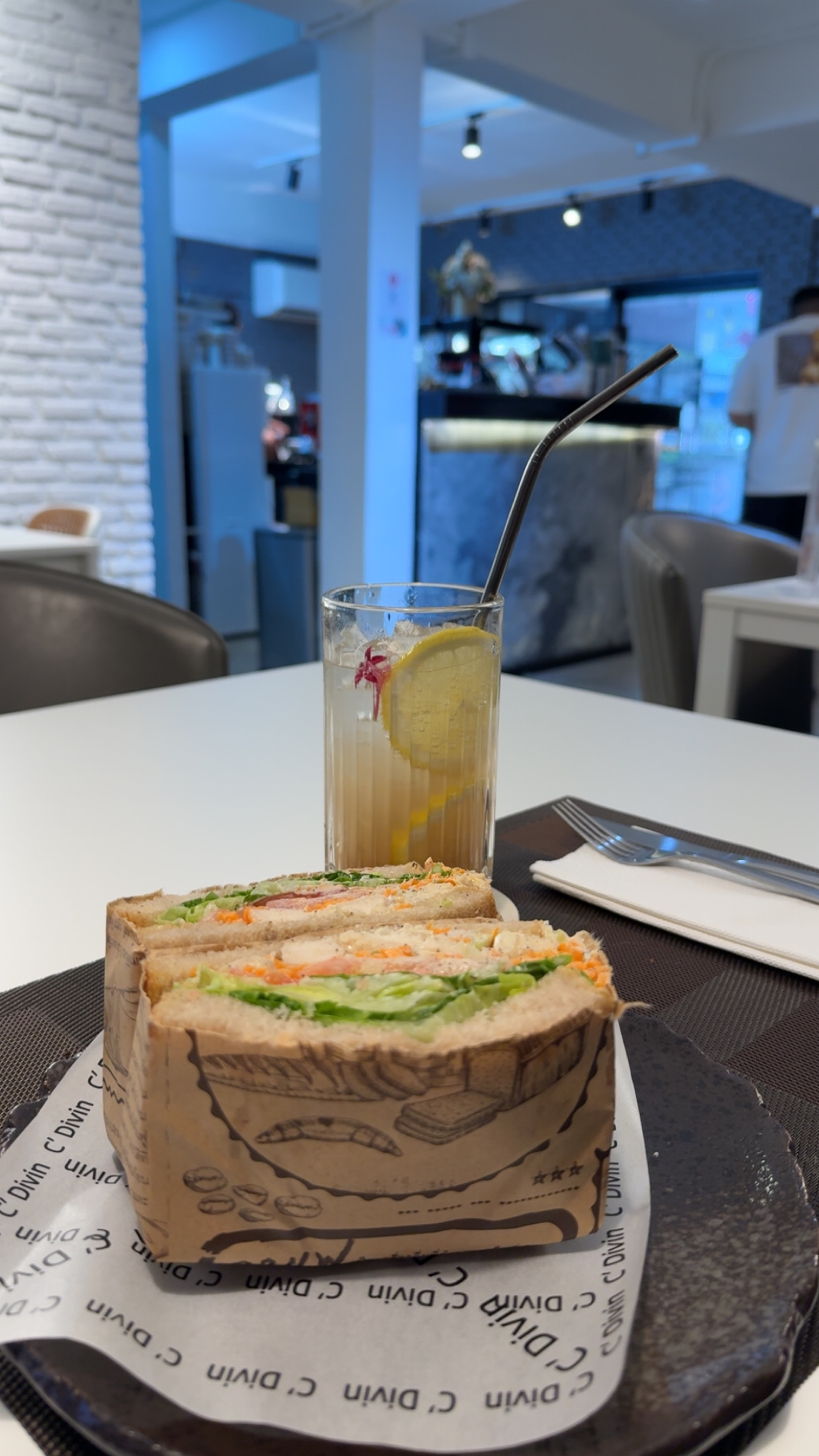 แซนด์วิชคลับไส้ผักและไก่ห่อกระดาษพร้อมน้ำมะนาวใส่แก้วบนถาดเสิร์ฟที่ร้าน C'Divin