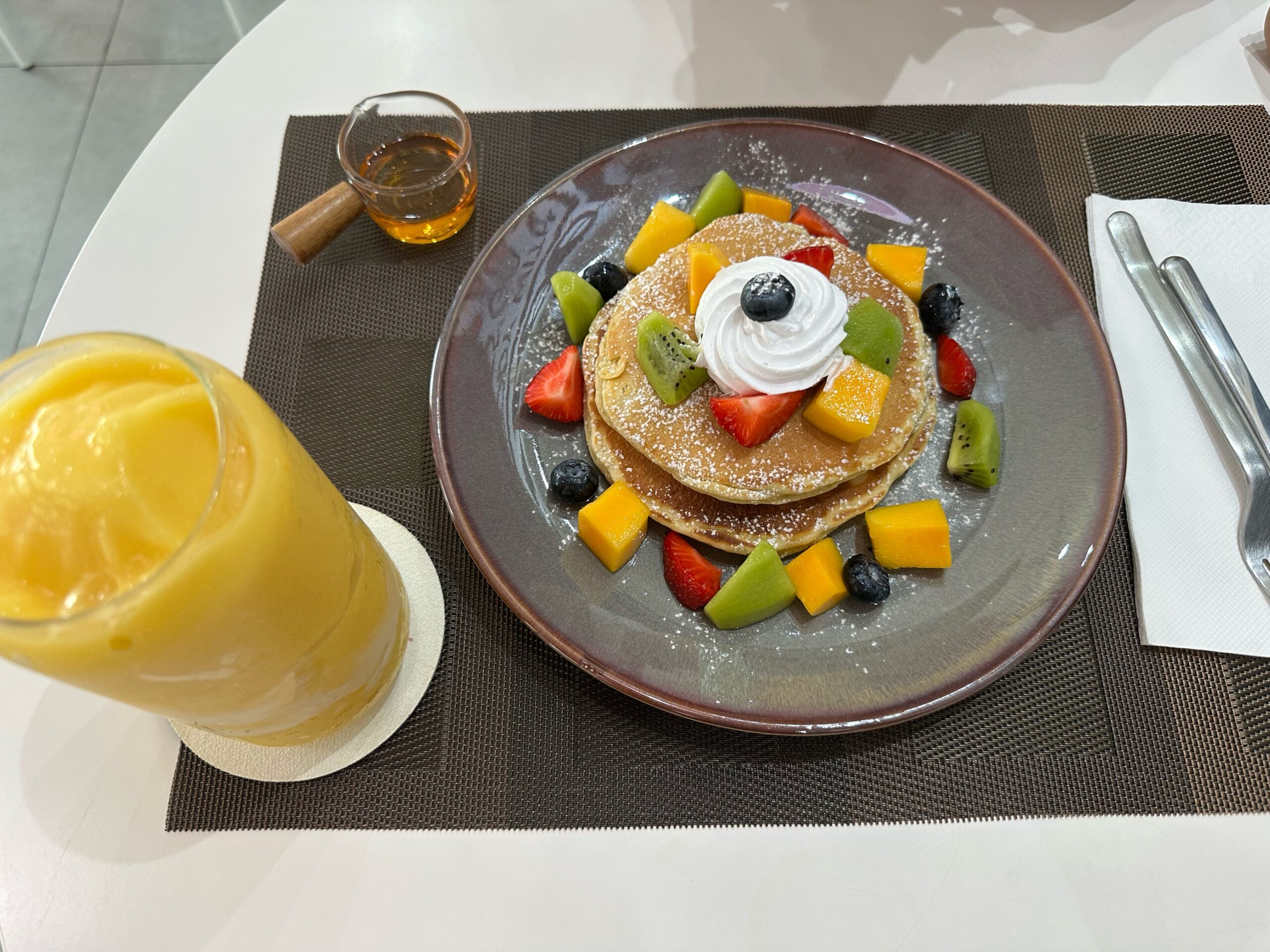 แพนเค้กซ้อนราดน้ำผึ้งตกแต่งด้วยผลไม้สด สตรอว์เบอร์รี่ กีวี มะม่วง บลูเบอร์รี่ วิปครีม พร้อมสมูทตี้มะม่วงเสิร์ฟบนจานสีเทาบนโต๊ะสีขาว