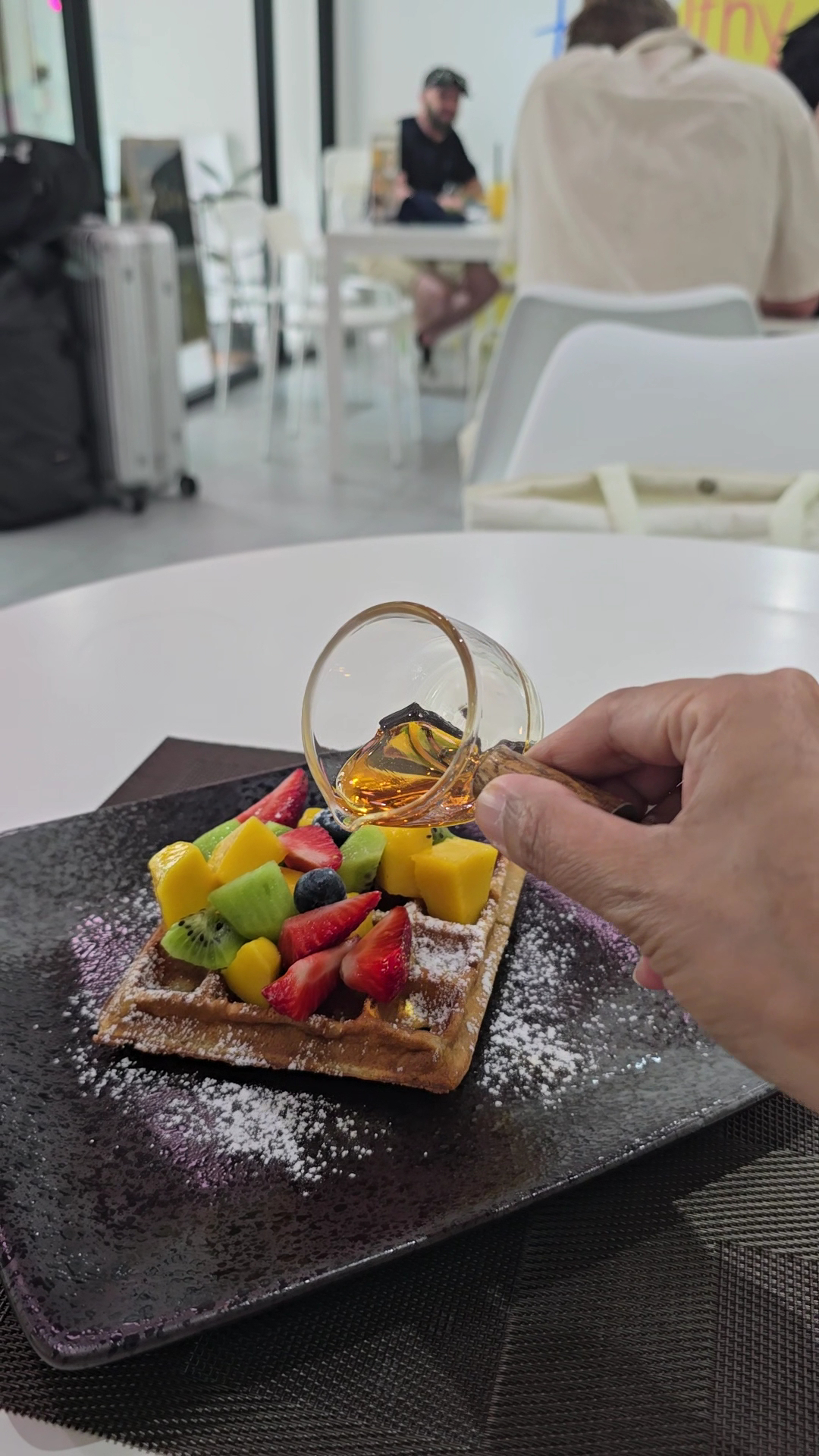 วาฟเฟิลท็อปปิ้งผลไม้สดหลากสีพร้อมน้ำผึ้งราดบนจานสีดำโรยผงน้ำตาลในร้านกาแฟบรรยากาศสบาย