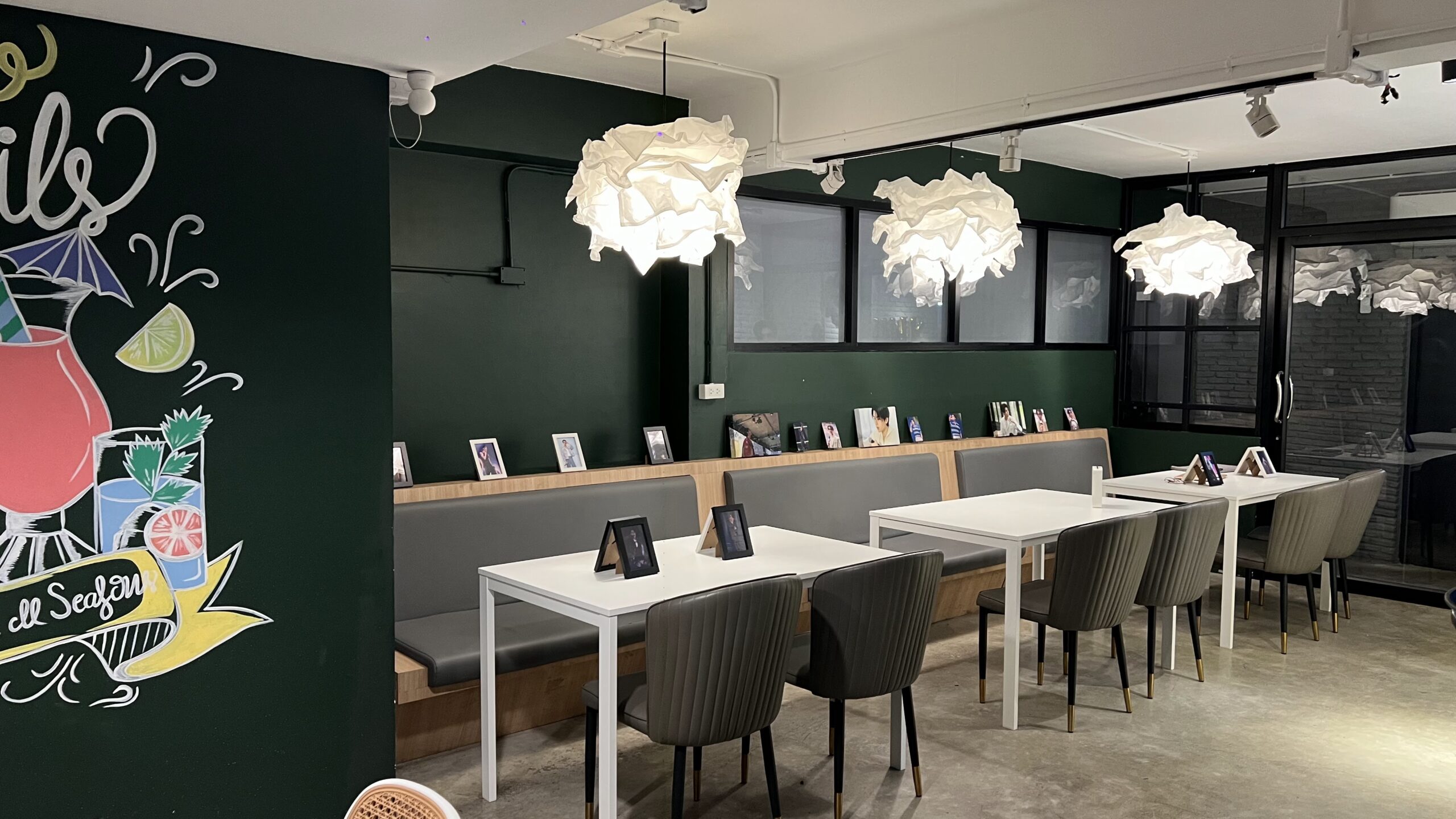 ภายในร้านคาเฟ่สไตล์โมเดิร์นผนังสีเขียวเข้มพร้อมโคมไฟสีขาวรูปทรงเมฆ โต๊ะสีขาวเก้าอี้สีเทาและภาพวาดค็อกเทลบนผนัง