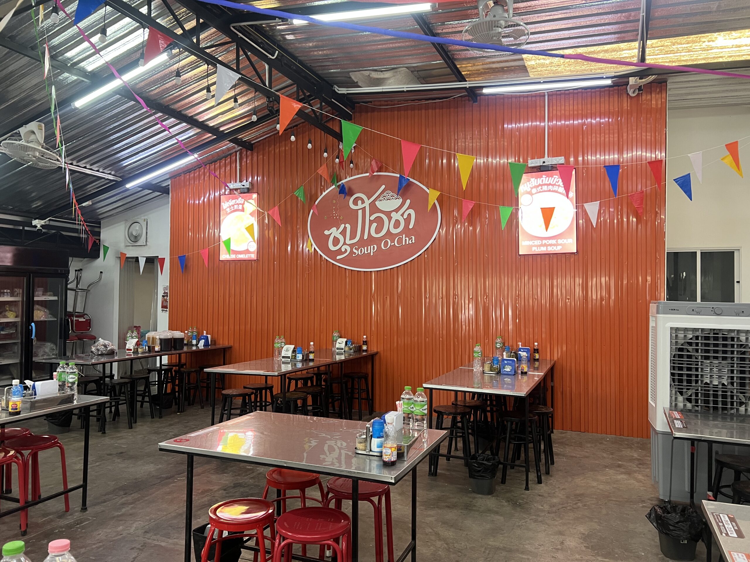ภายในร้านซุปโอชา บรรยากาศแบบเปิดโล่งมีโต๊ะเก้าอี้โลหะ ผนังสังกะสีสีส้มพร้อมป้ายชื่อร้านและธงสามเหลี่ยมหลากสีตกแต่ง