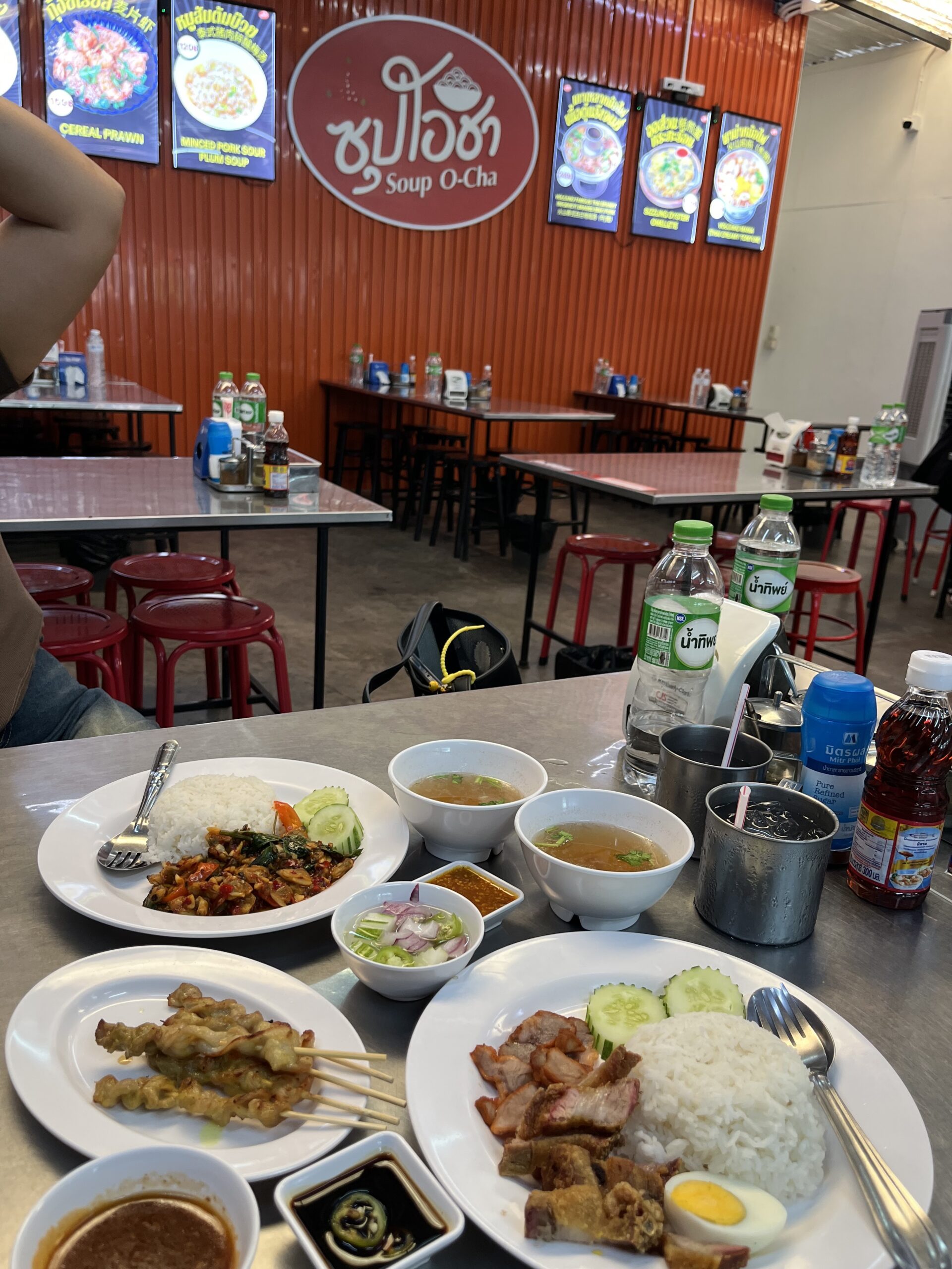อาหารไทยหลายจานที่ร้านซุปโอชา มีข้าวราดกะเพรา หมูแดงข้าวมันไข่ต้ม สะเต๊ะ ซุป และน้ำจิ้ม บนโต๊ะสแตนเลสพร้อมเครื่องดื่ม