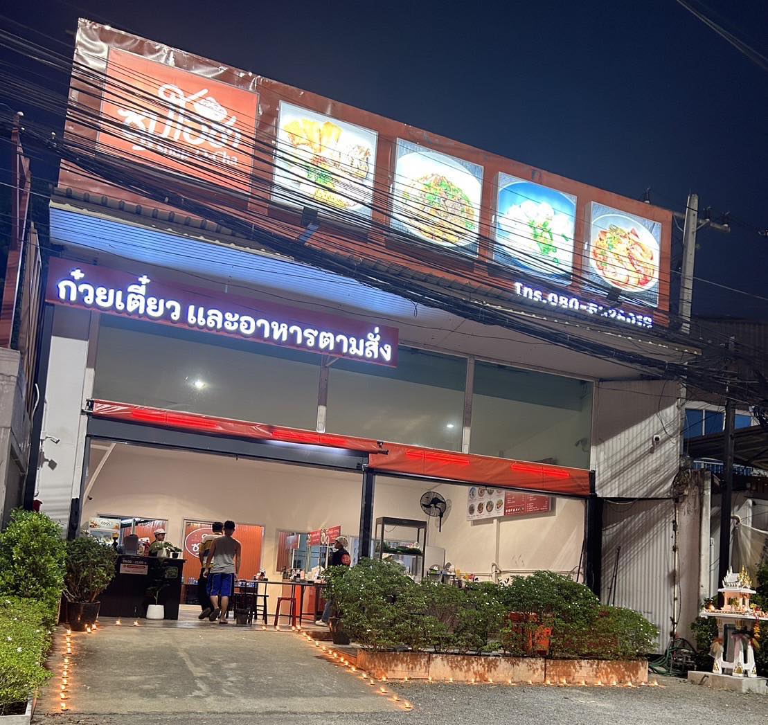 ร้านก๋วยเตี๋ยวและอาหารตามสั่งเปิดไฟสว่างในเวลากลางคืน มีป้ายไฟสีแดงและรูปอาหารประดับด้านบน
