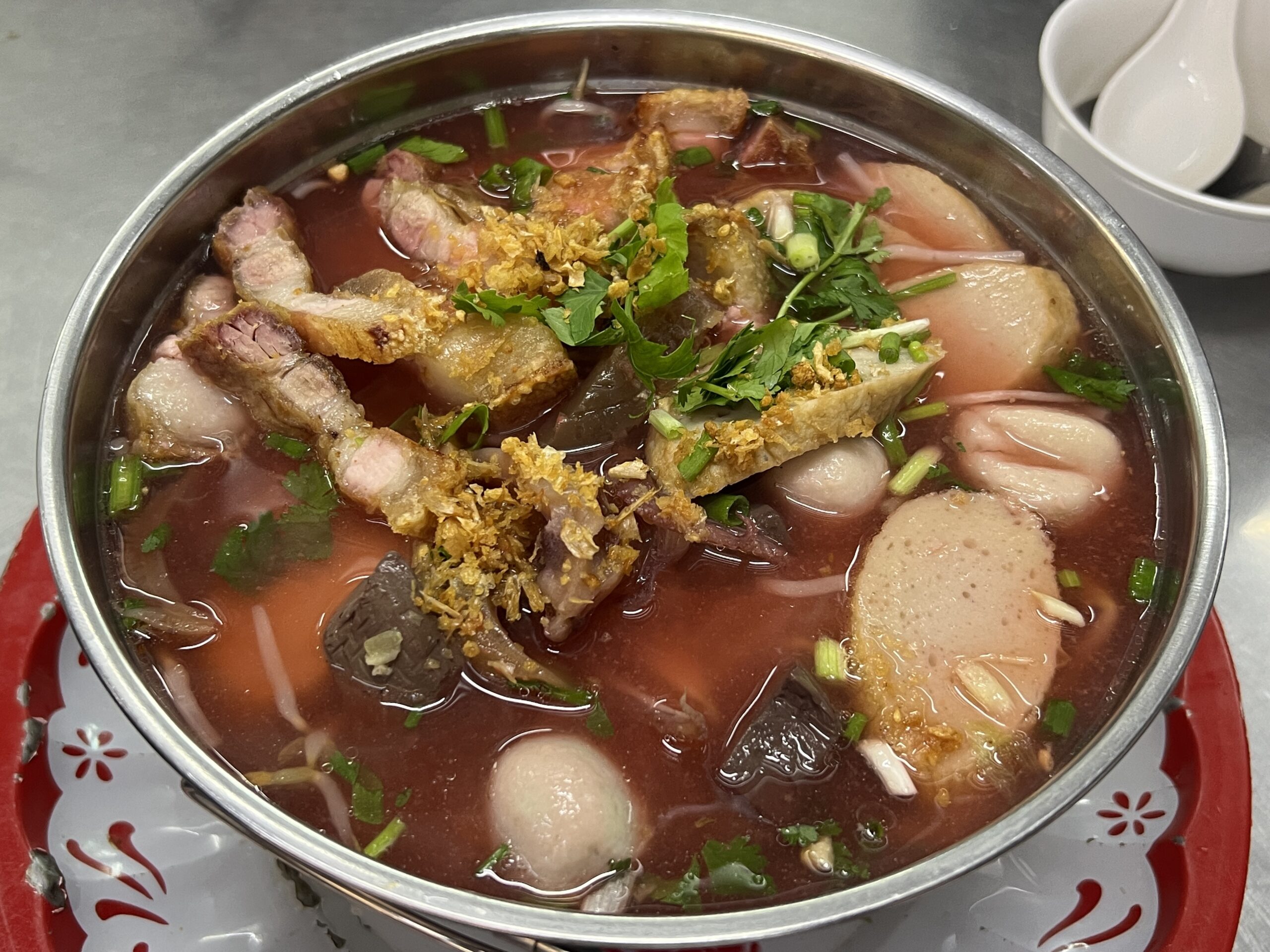 ชามเย็นตาโฟน้ำแดงขนาดใหญ่ใส่หมูกรอบ ลูกชิ้น เลือดหมู เต้าหู้ทอด โรยกระเทียมเจียวและผักชี เสิร์ฟในชามสแตนเลสบนถาดแดง