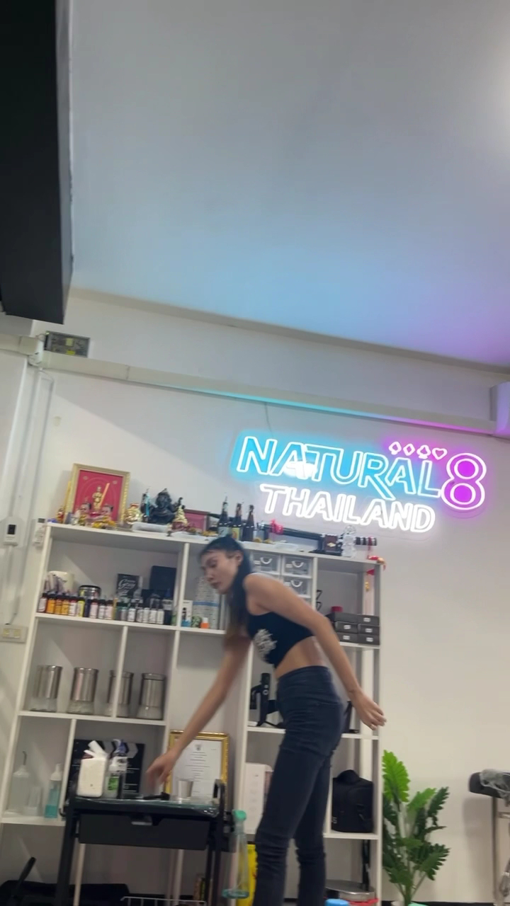 พนักงานสาวในชุดดำกำลังทำงานหน้าป้ายไฟนีออน Natural8 Thailand พร้อมชั้นวางของและเครื่องดื่ม