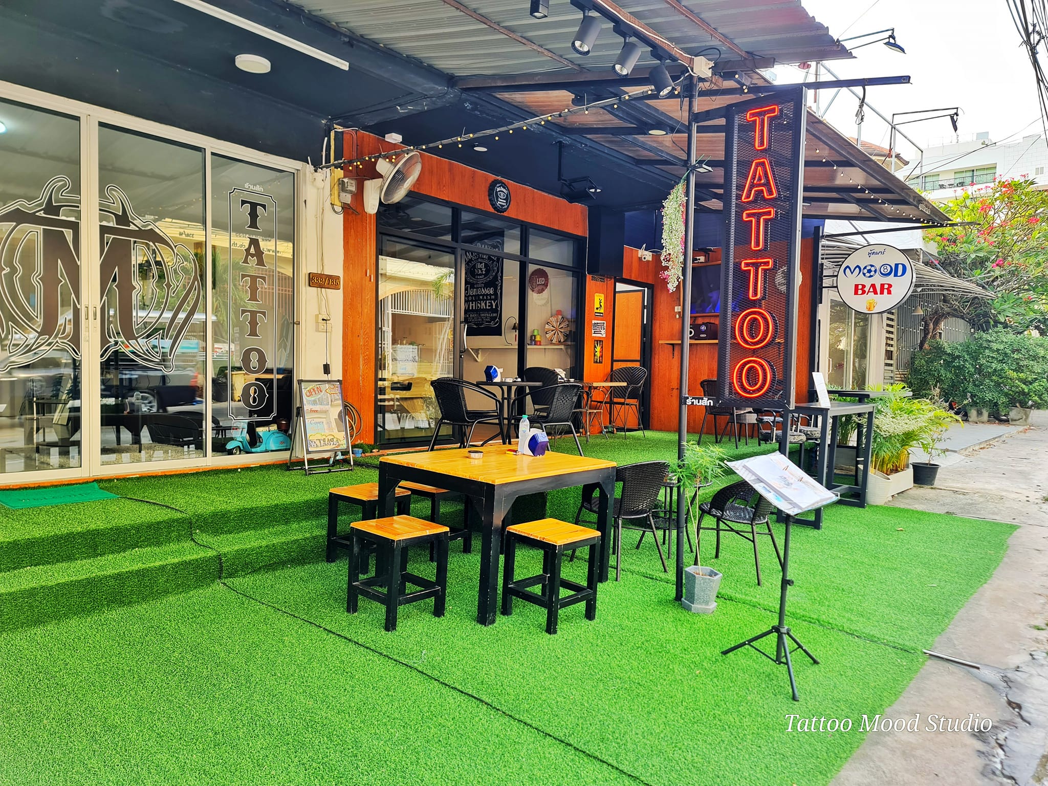 ร้าน Tattoo Mood Studio พร้อมบาร์ Mood Bar มีโต๊ะเก้าอี้นั่งด้านนอกบนหญ้าเทียมสีเขียว ป้ายไฟนีออนสีแดงเขียนว่า TATTOO และป้าย Mood Bar
