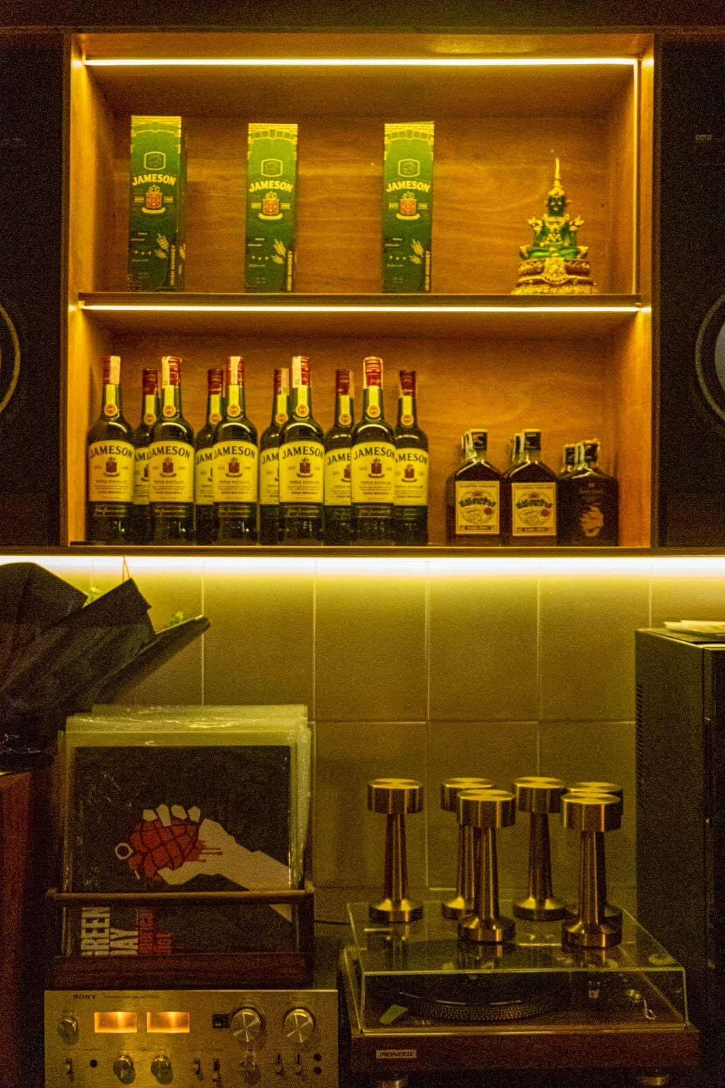 ชั้นวางขวดวิสกี้ Jameson เรียงแถวพร้อมกล่อง มีพระพุทธรูปตกแต่ง เครื่องเล่นแผ่นเสียงและแอมป์ Sony ด้านล่างในบาร์บรรยากาศอบอุ่น