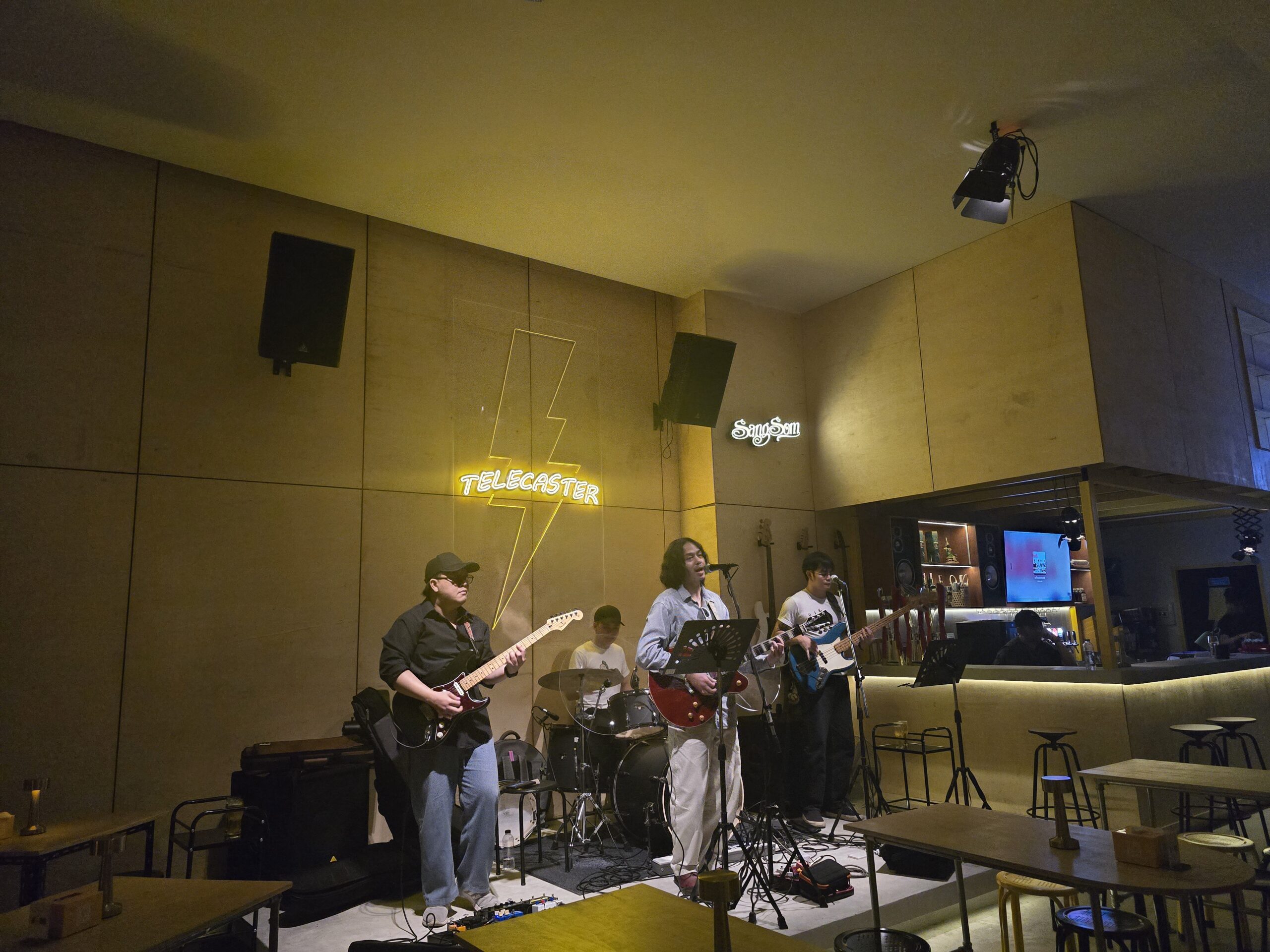 วงดนตรีสดกำลังเล่นบนเวทีในบาร์ Telecaster พร้อมป้ายไฟนีออนรูปสายฟ้าและนักดนตรีหลายคนเล่นกีตาร์และกลอง