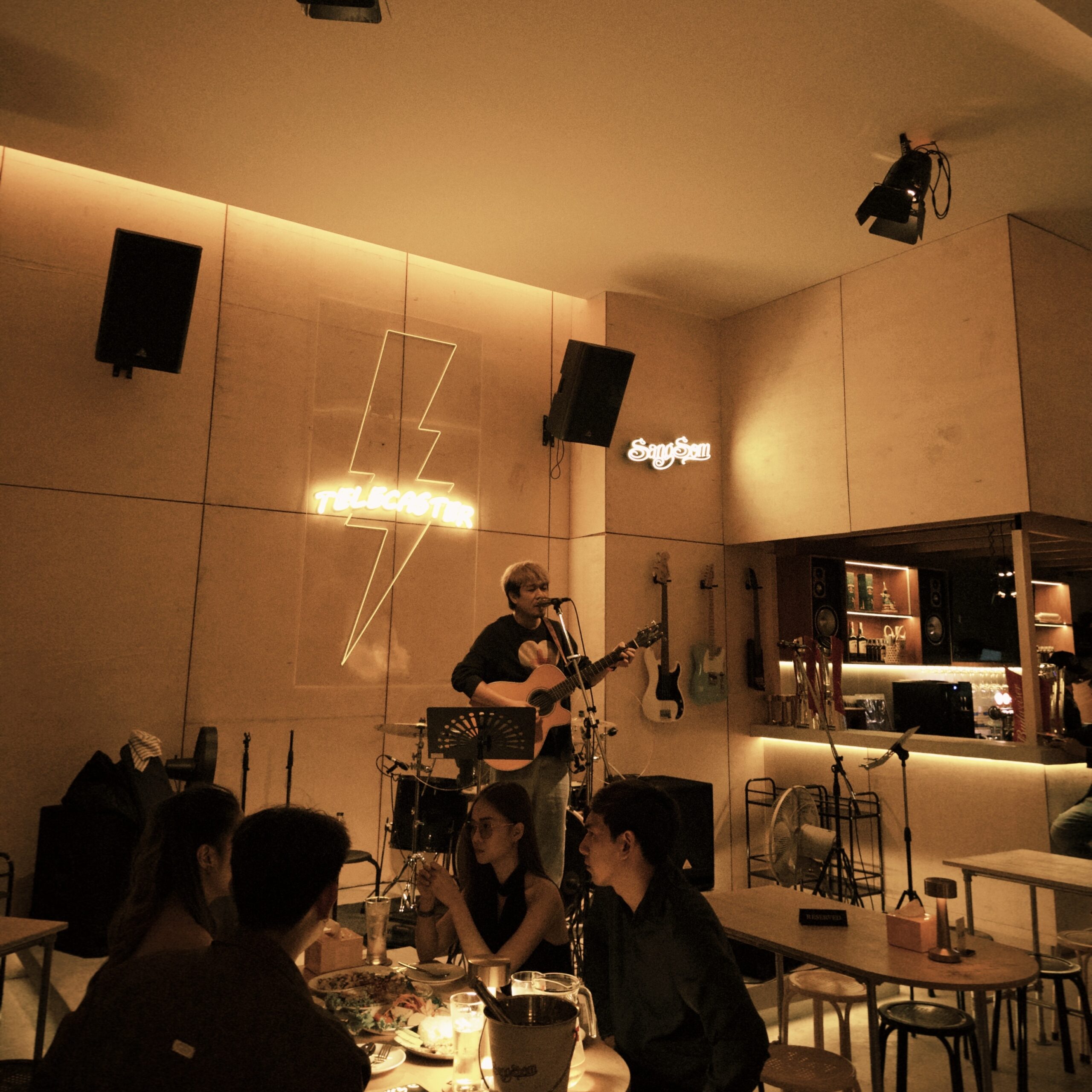 นักดนตรีเล่นกีตาร์โปร่งร้องเพลงสดในบาร์ Telecaster บรรยากาศอบอุ่นมีไฟนีออนรูปสายฟ้าและลูกค้านั่งทานอาหารฟังเพลง