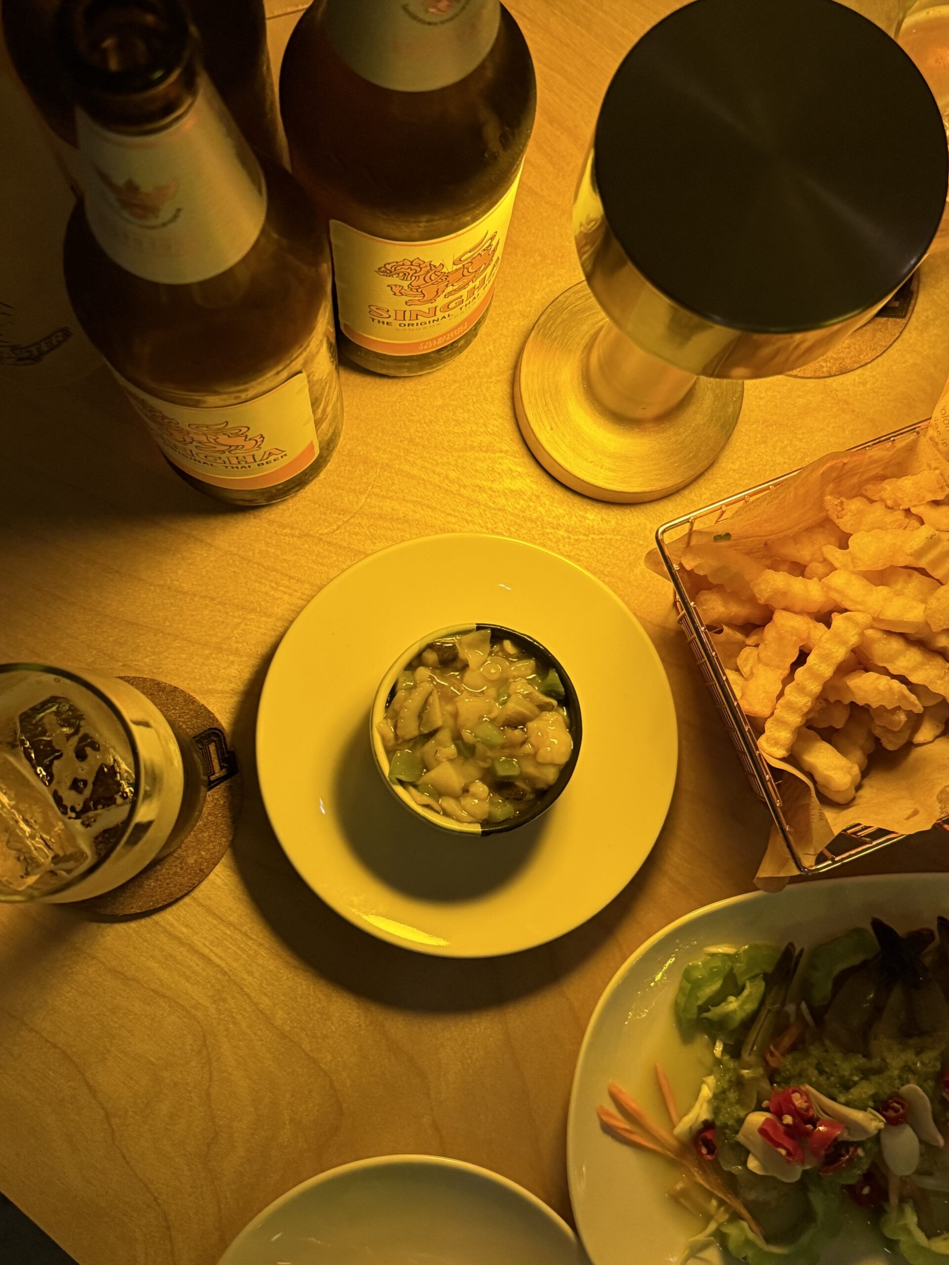 โต๊ะอาหารไทยกับเบียร์สิงห์สองขวด จานน้ำจิ้ม เฟรนช์ฟรายส์ และสลัดผักสด บรรยากาศร้านอาหารแสงสีอบอุ่น