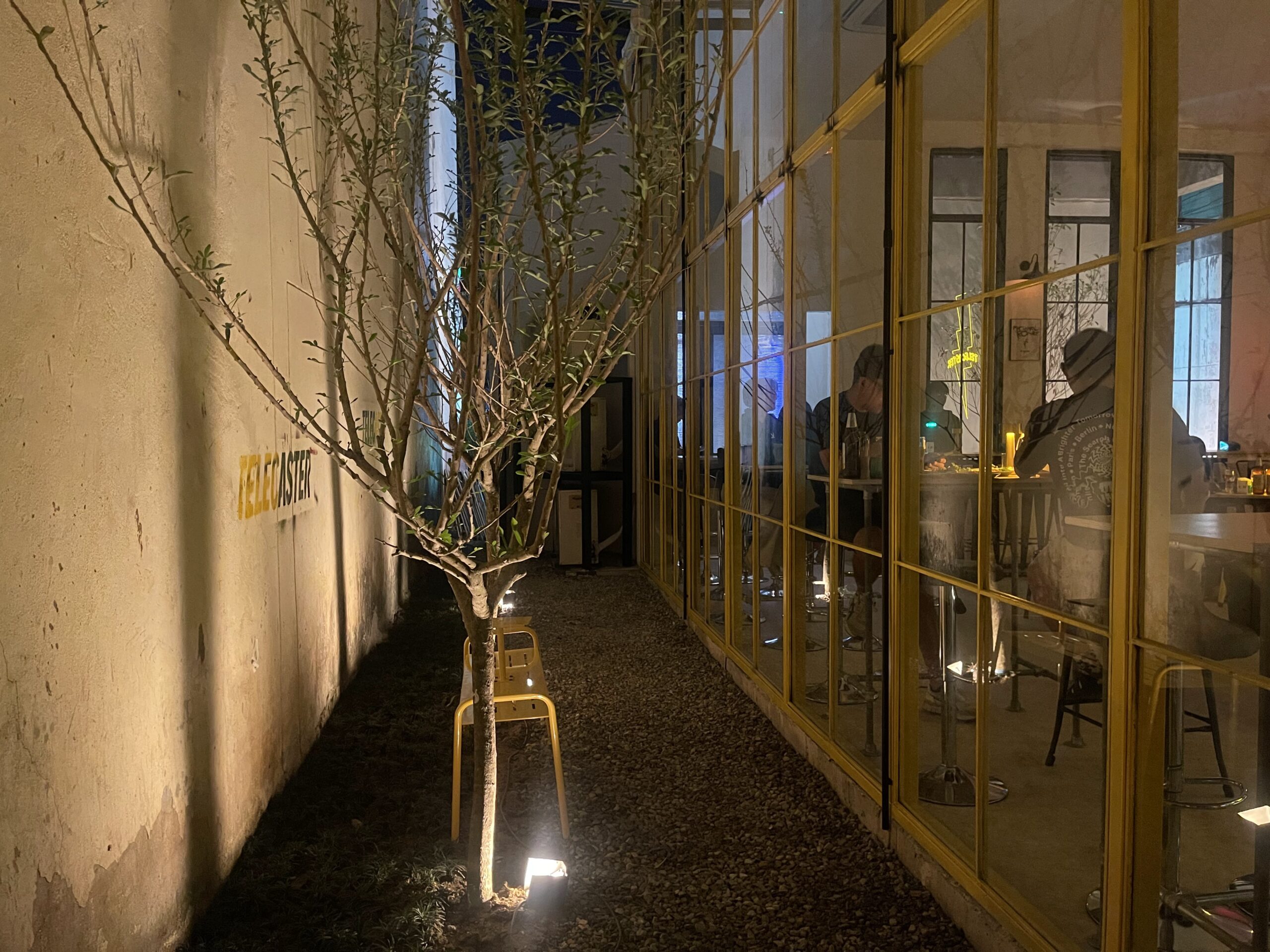 ทางเดินด้านข้างร้าน Telecaster ในยามค่ำคืน มีต้นไม้ประดับไฟส่องสว่าง เก้าอี้สีเหลือง และกระจกบานใหญ่กรอบสีทองมองเห็นบรรยากาศภายในร้าน
