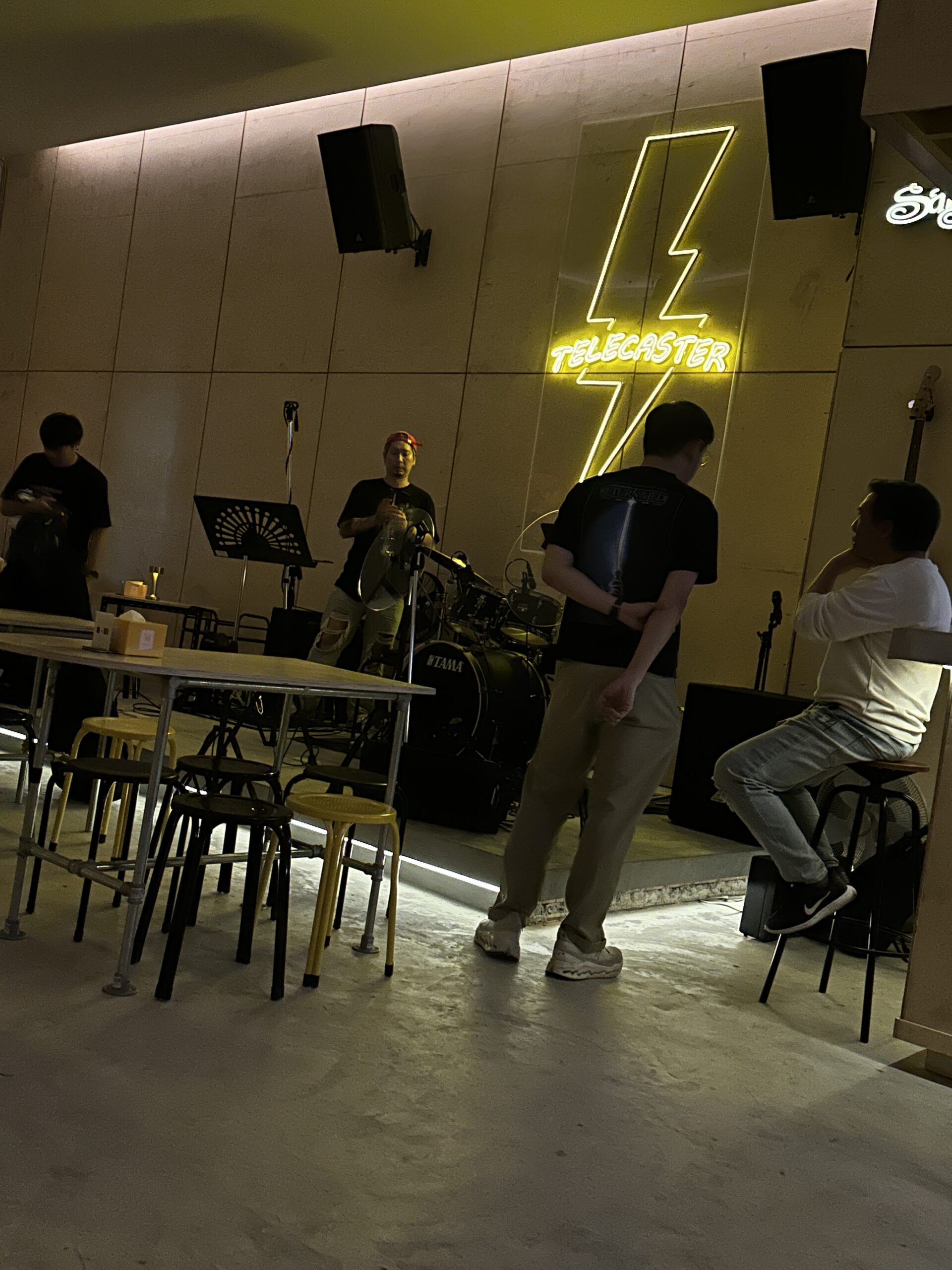 บรรยากาศภายในร้าน Telecaster บาร์ดนตรีสดพร้อมเวทีกลองชุด TAMA ป้ายไฟนีออนรูปสายฟ้าสีเหลือง และลูกค้านั่งพักผ่อน