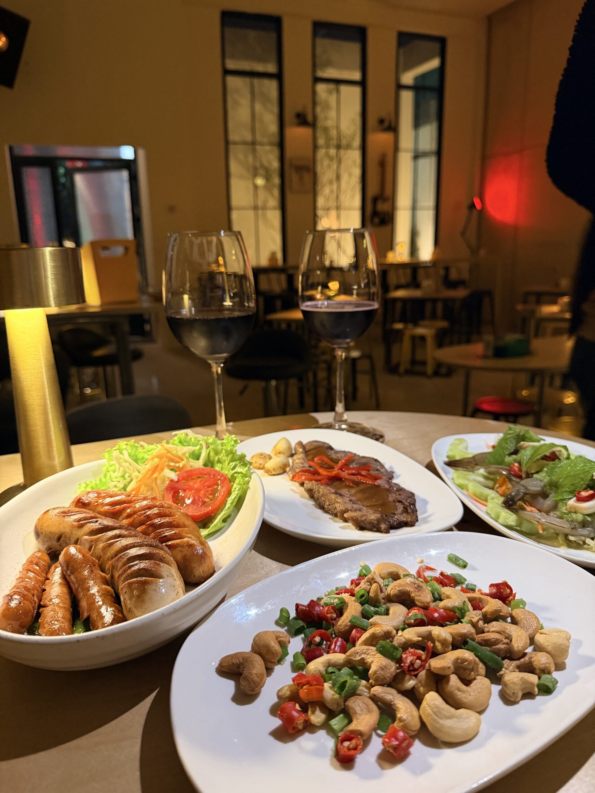 โต๊ะอาหารในร้านอาหารพร้อมไวน์แดง 2 แก้ว จานไส้กรอกย่างกับสลัด เนื้อสเต็ก เม็ดมะม่วงหิมพานต์ผัดพริก และสลัดกุ้งสด