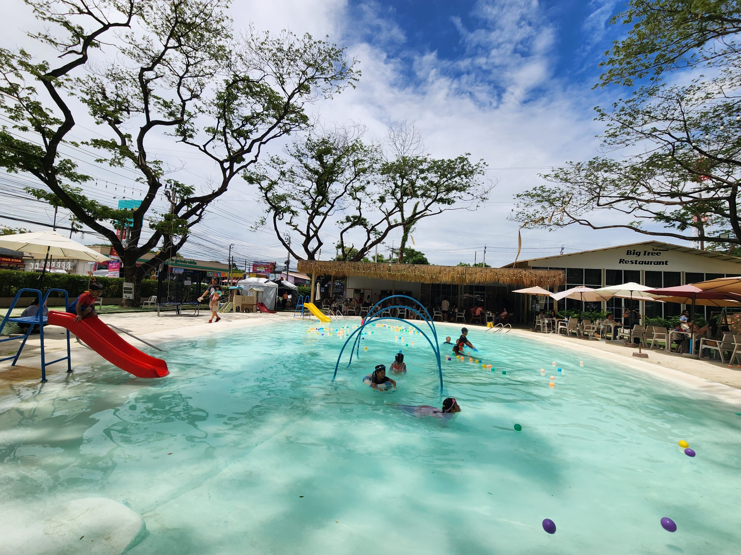 ร้านอาหาร Big Tree Restaurant พร้อมสระว่ายน้ำสำหรับเด็กที่มีสไลเดอร์สีแดงและเครื่องเล่นน้ำ ล้อมรอบด้วยต้นไม้ใหญ่และร่มกันแดด