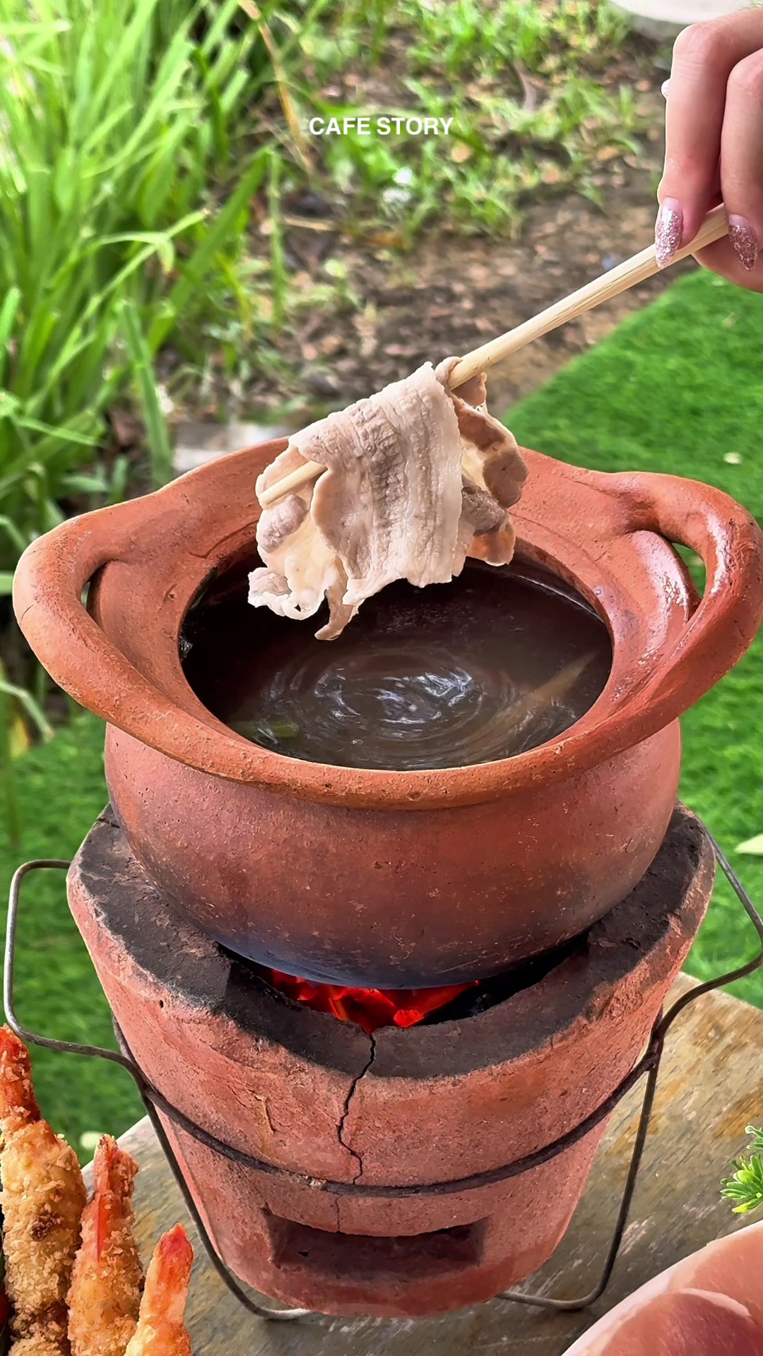 หม้อดินเผาตั้งบนเตาถ่านแดง มีคีบเนื้อหมูสไลซ์ด้วยตะเกียบจุ่มในน้ำซุปร้อน สไตล์ชาบูหม้อดิน