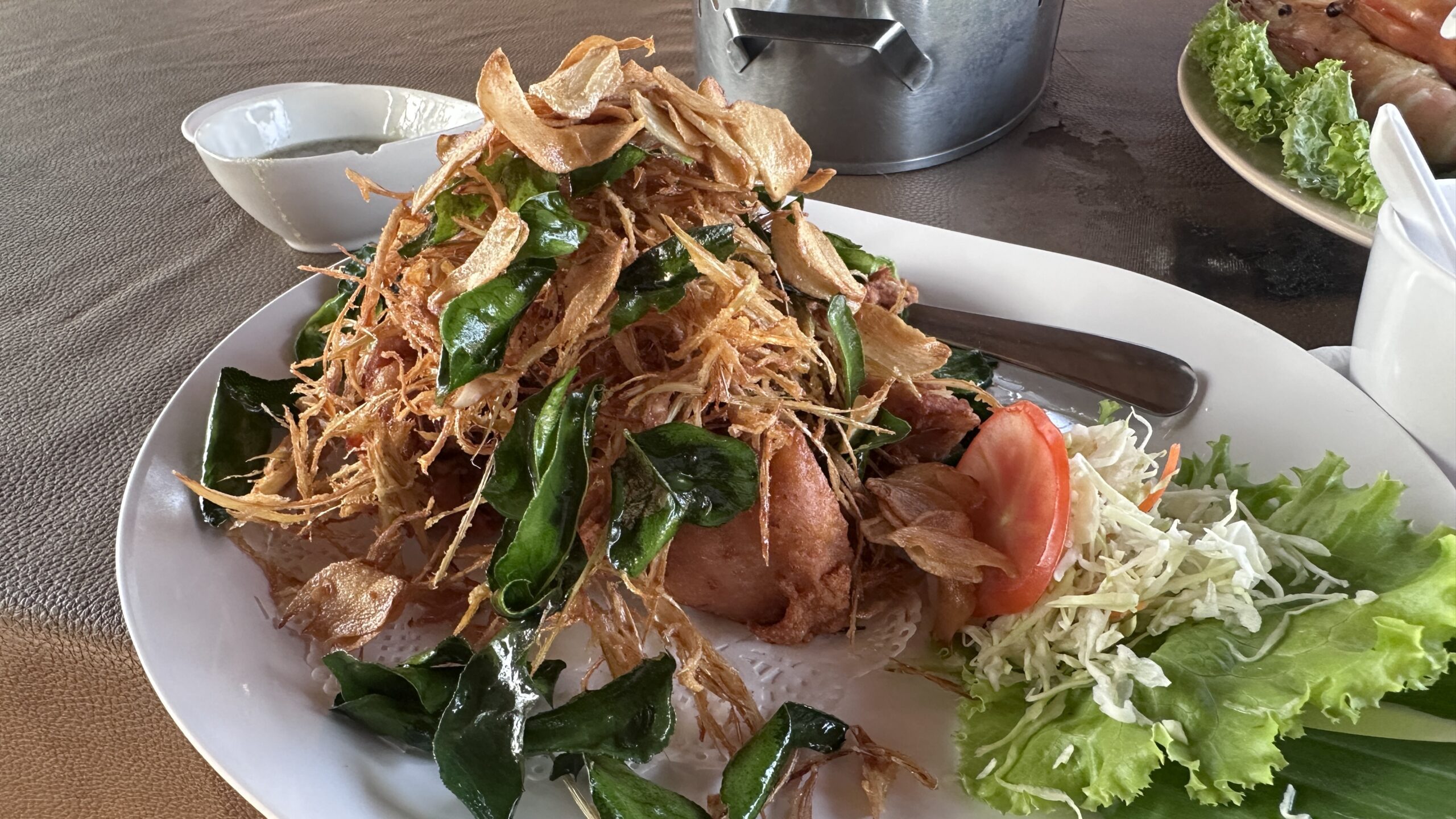 ปลาทอดกรอบราดสมุนไพร ตกแต่งด้วยใบกะเพราทอด กระเทียมเจียว และผักสลัด บนจานขาวในร้านอาหาร