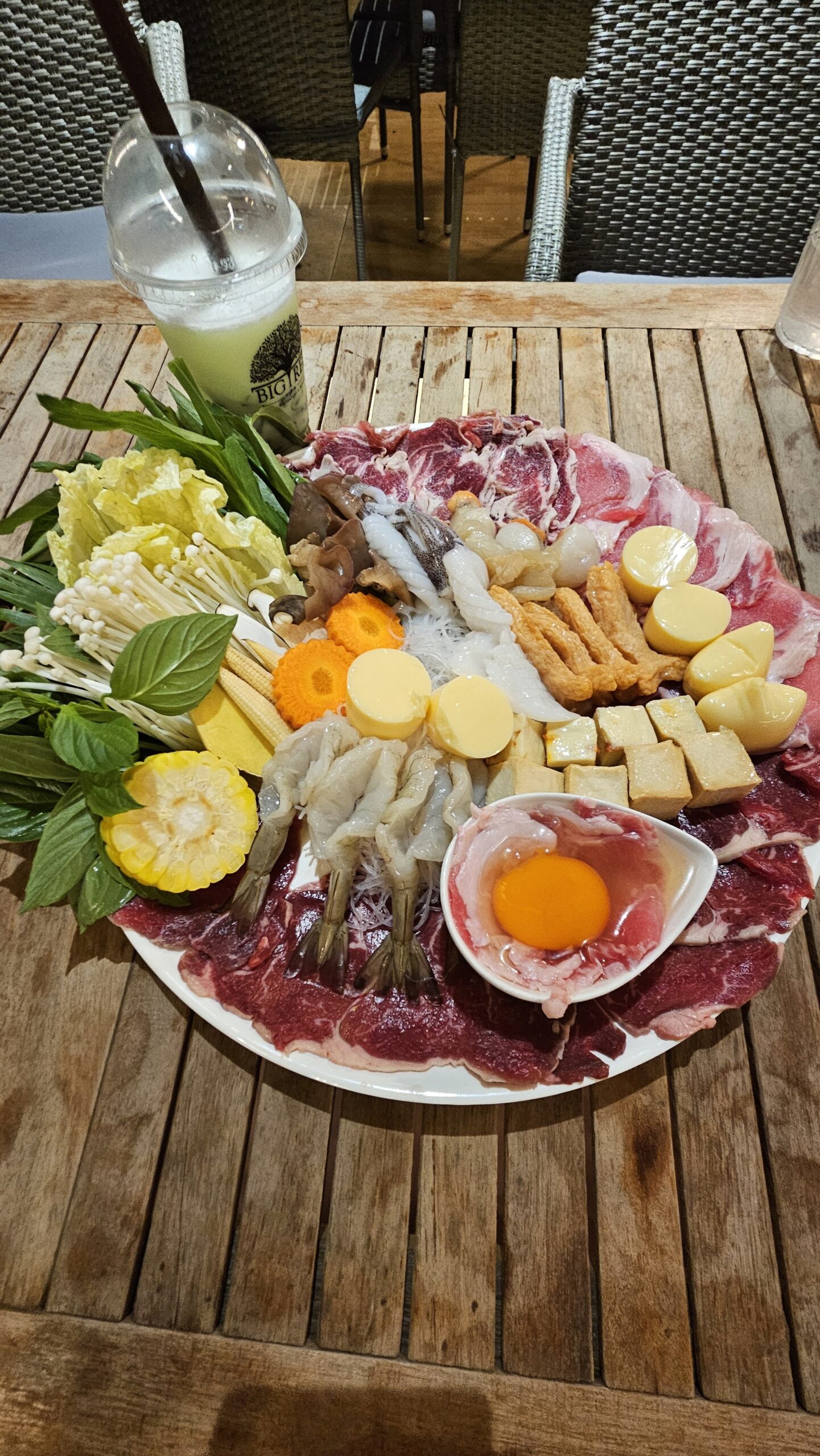 จานชาบูขนาดใหญ่บนโต๊ะไม้ มีเนื้อวัวสไลด์ กุ้งสด เห็ดเข็มทอง ข้าวโพดอ่อน แครอท เต้าหู้ ไข่แดงดิบ ผักบุ้ง โหระพา และเครื่องดื่มแก้วพลาสติก