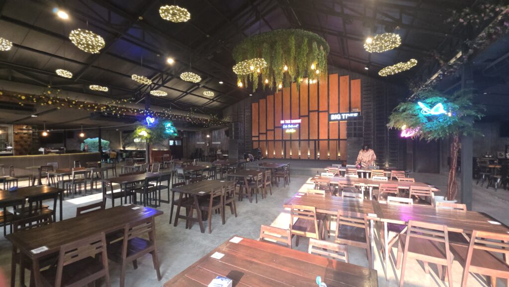 ภายในร้านอาหาร Big Tree ตกแต่งสวยงามด้วยโคมไฟกลมเรืองแสง ต้นไม้ประดับห้อยจากเพดาน โต๊ะเก้าอี้ไม้จัดเรียงเป็นระเบียบในห้องโถงกว้างใหญ่