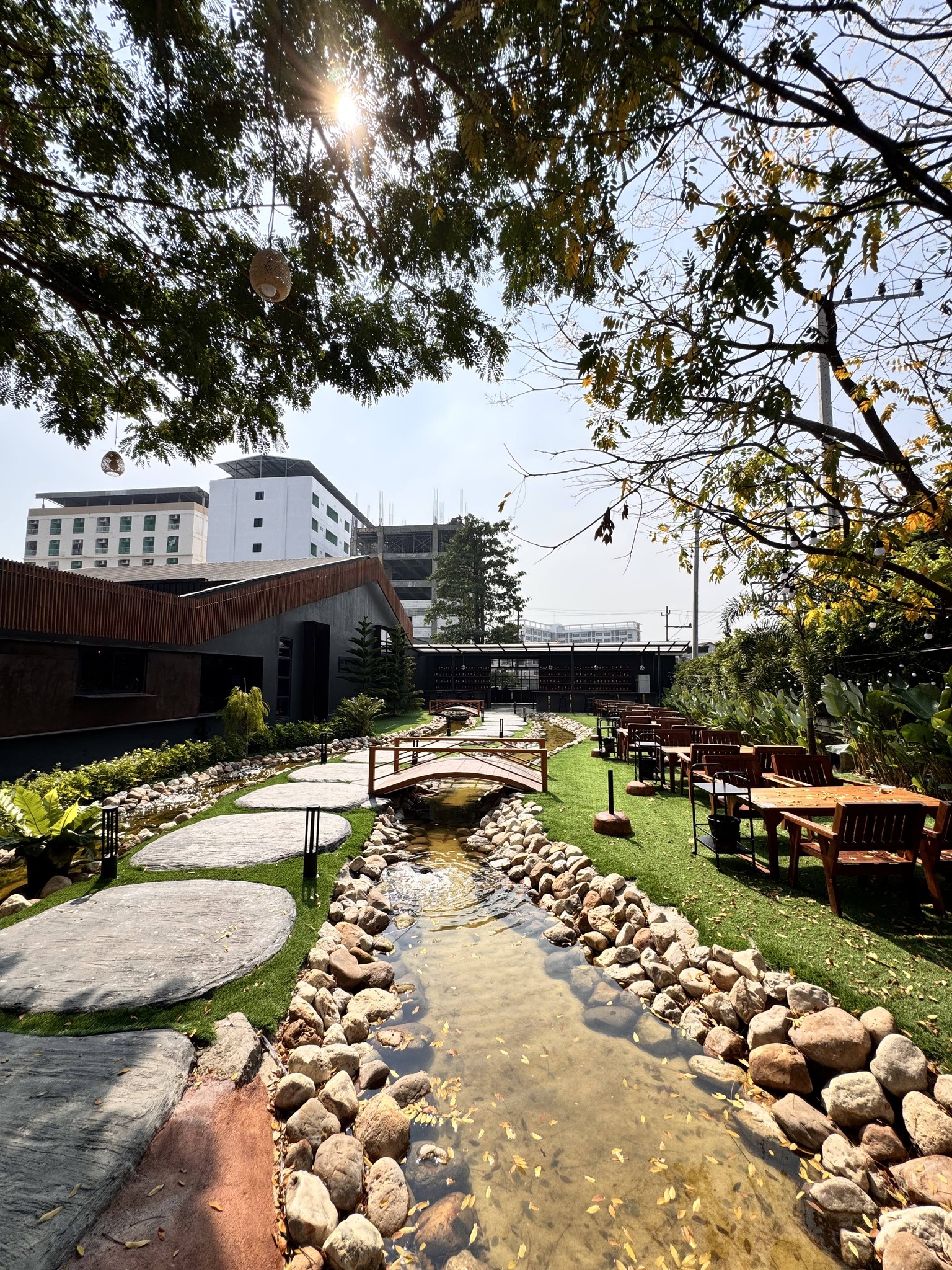 บรรยากาศร้านอาหารกลางแจ้งพร้อมลำธารหินธรรมชาติ สะพานไม้ สนามหญ้าสีเขียว และโต๊ะอาหารไม้ ใต้ร่มไม้ใหญ่ในยามแดดอ่อน