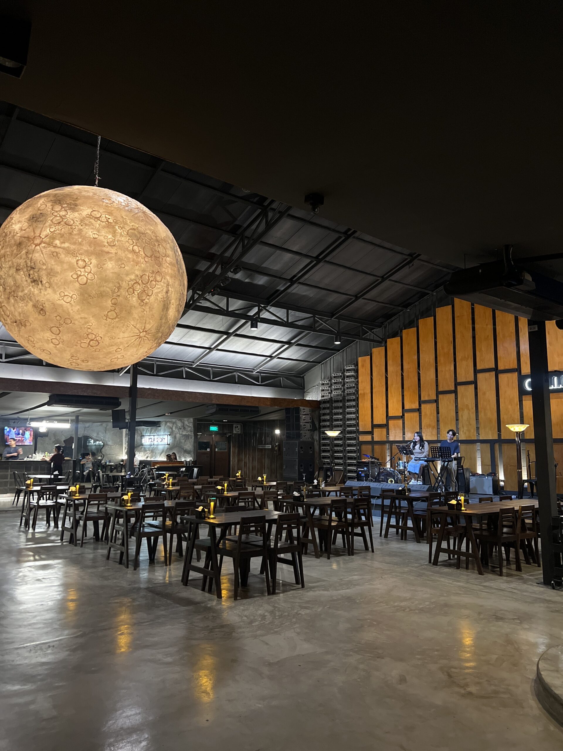 ร้านอาหารสไตล์อินดัสเทรียลกว้างขวาง มีโคมไฟรูปดวงจันทร์ขนาดใหญ่แขวนจากเพดาน โต๊ะไม้เรียงเป็นแถว และเวทีดนตรีสดพร้อมนักร้อง