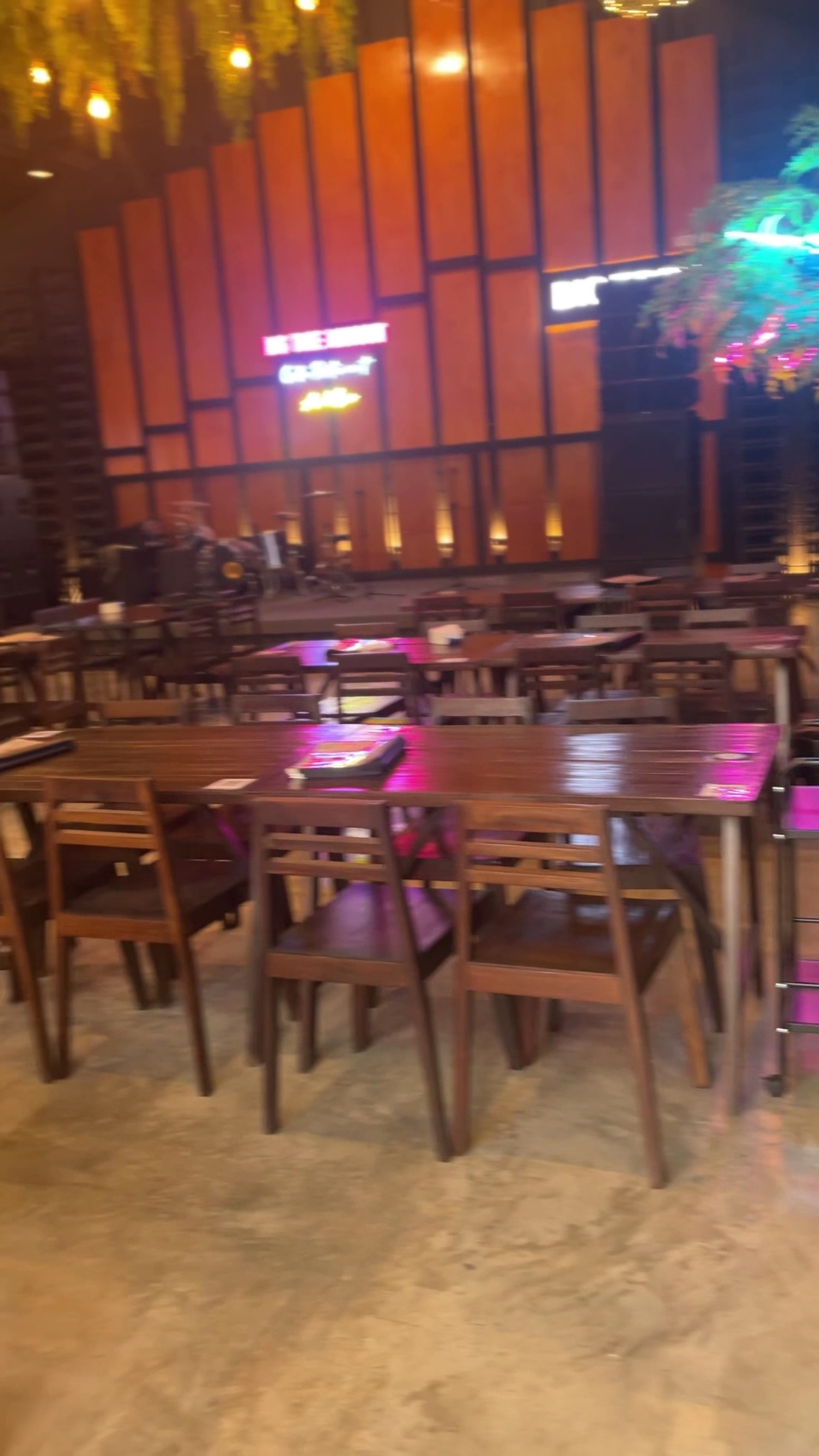 บรรยากาศภายในร้านอาหารที่มีโต๊ะไม้เก้าอี้ไม้จัดเรียงเป็นระเบียบ พร้อมเวทีดนตรีสดด้านหลังตกแต่งด้วยผนังไม้และไฟนีออนสีสันสดใส