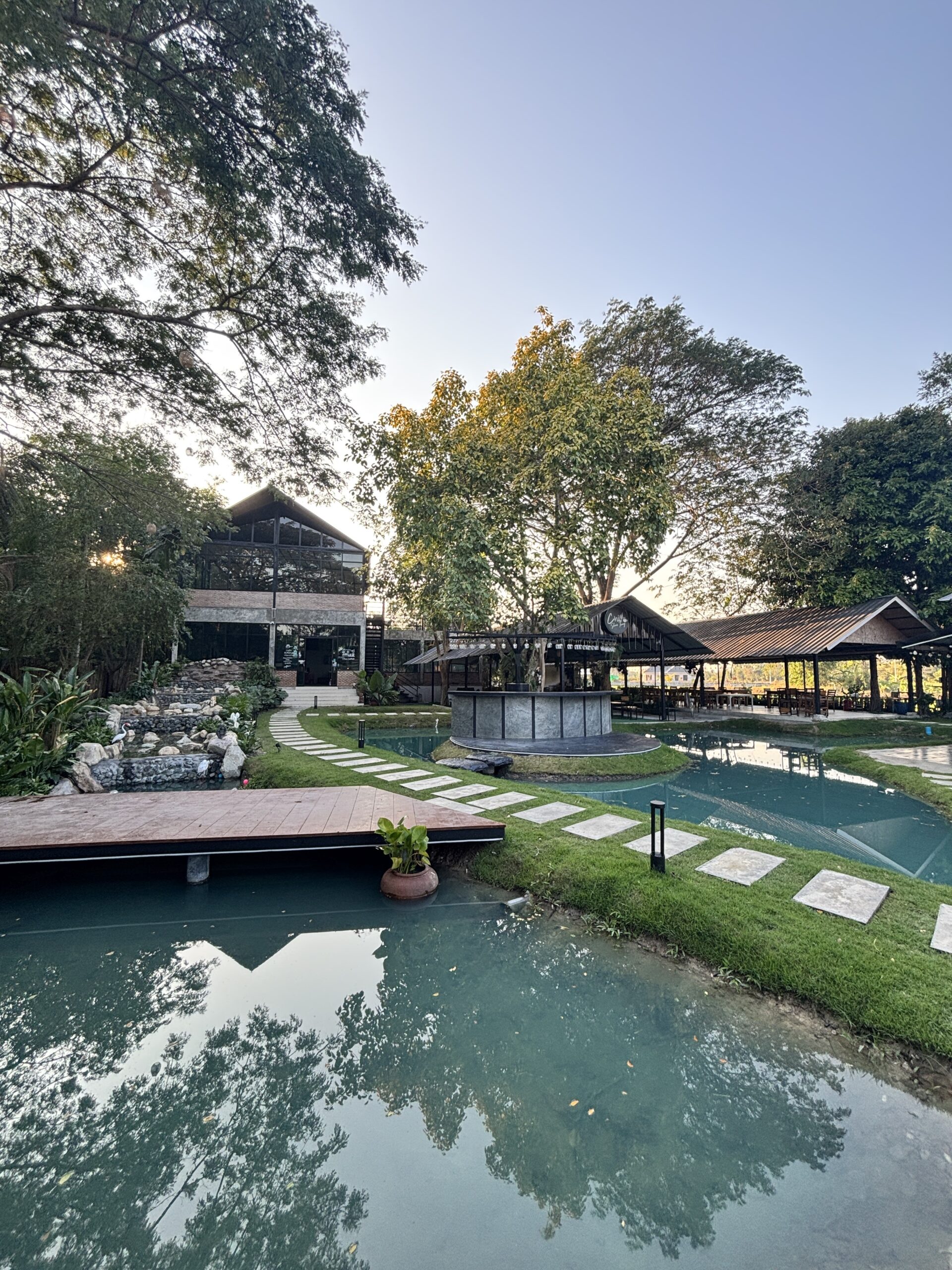 ร้านอาหารริมน้ำพร้อมศาลาและสระน้ำสีเขียวล้อมรอบด้วยต้นไม้ใหญ่ มีทางเดินหินและระเบียงไม้
