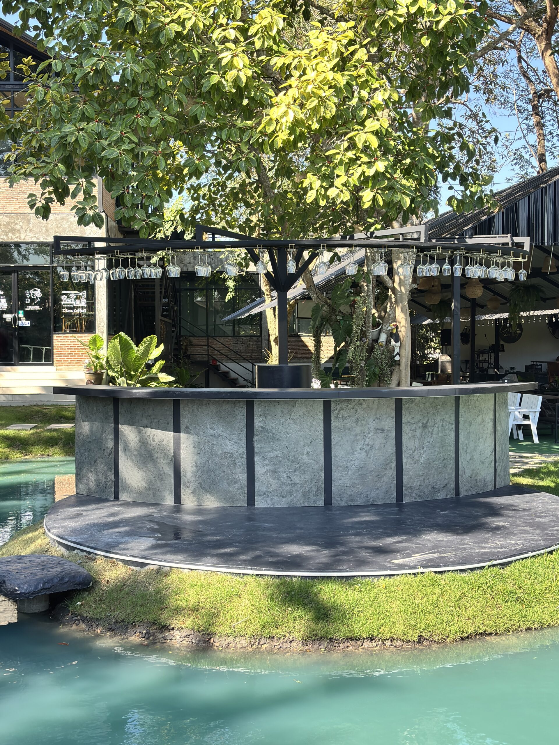 บาร์เคาน์เตอร์กลางแจ้งรูปโค้งทำจากหินอ่อนสีเทา ล้อมรอบด้วยสระน้ำสีเขียวมรกตและสวนร่มรื่นใต้ต้นไม้ใหญ่ มีแก้วไวน์แขวนเรียงรายบนโครงเหล็กด้านบน