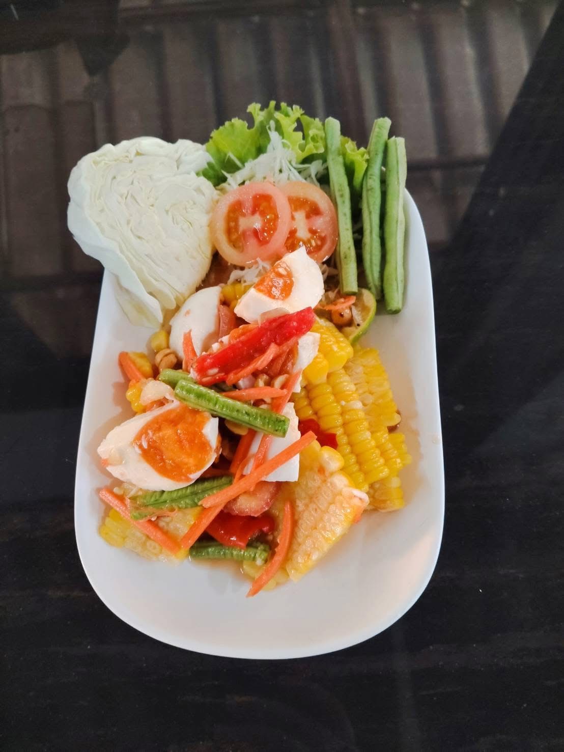 ส้มตำข้าวโพดไข่เค็มพร้อมผักสดกะหล่ำปลี ถั่วฝักยาว และมะเขือเทศ บนจานขาว