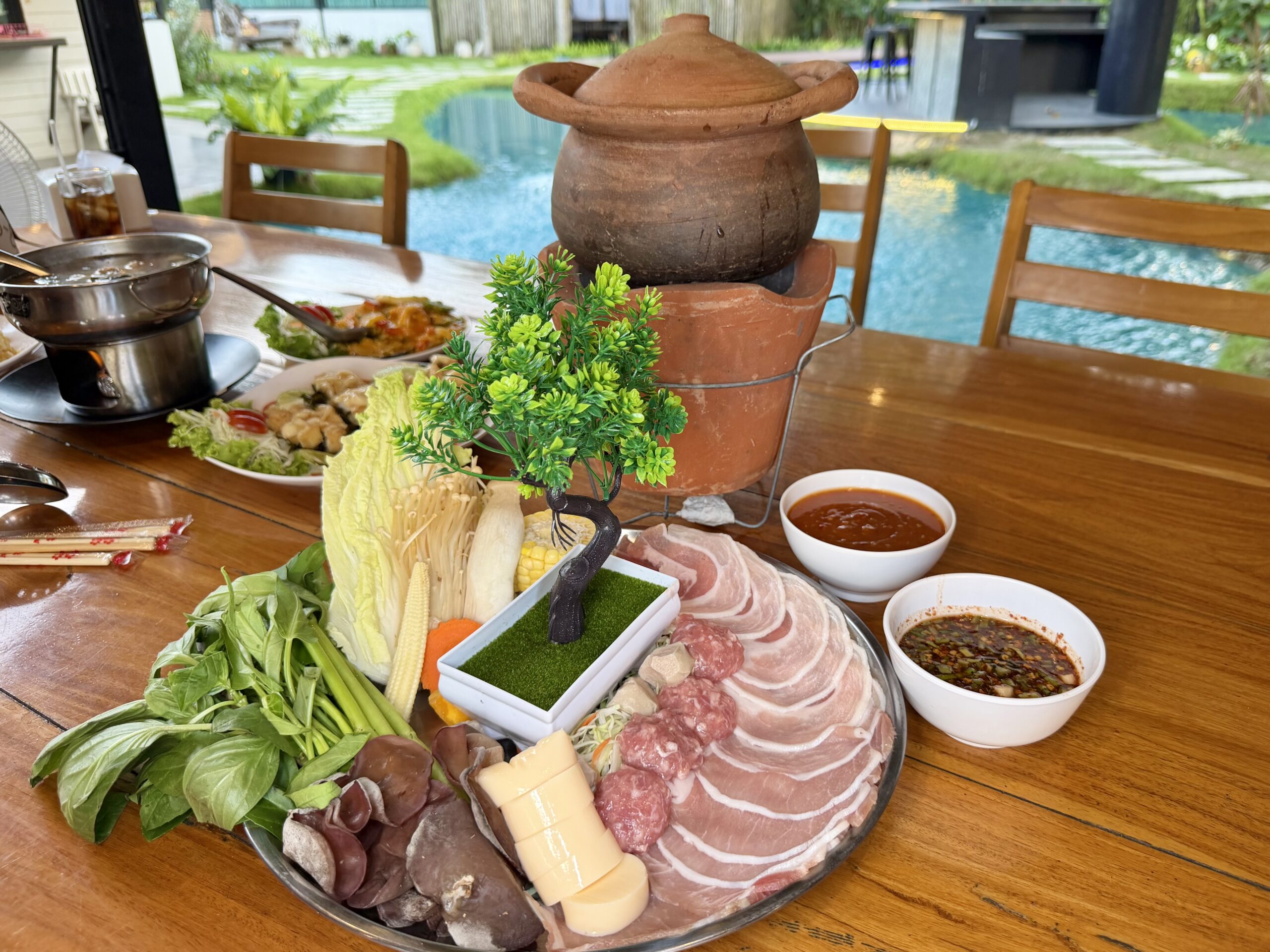 จิ้มจุ่มหม้อดินพร้อมจานเนื้อหมูสไลด์ ผักสด เห็ด ข้าวโพดอ่อน และน้ำจิ้ม บนโต๊ะไม้ริมสระน้ำ