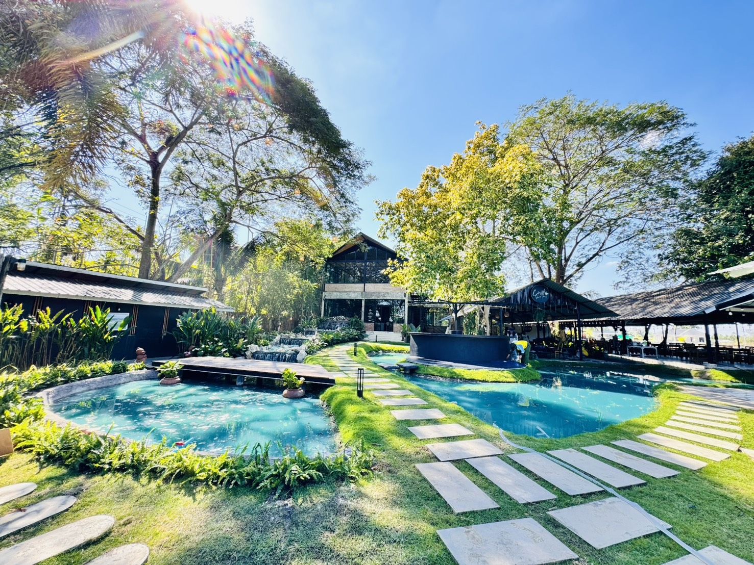 บรรยากาศร้านอาหารสไตล์คาเฟ่กลางสวนเขียวขจี มีสระน้ำสีฟ้าใส ทางเดินหินปูน ล้อมรอบด้วยต้นไม้ใหญ่ในวันแดดสดใส