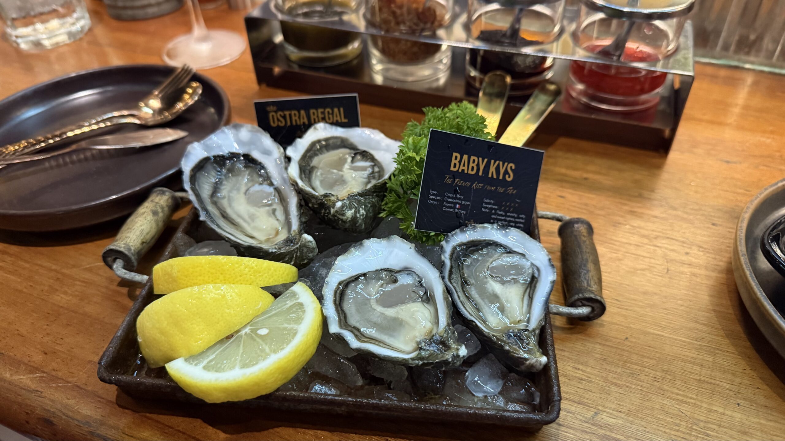 หอยนางรมสด Baby Kys จำนวน 5 ตัว เสิร์ฟบนถาดน้ำแข็งพร้อมเลมอนและผักชีฝรั่ง บนโต๊ะไม้ในร้านอาหาร
