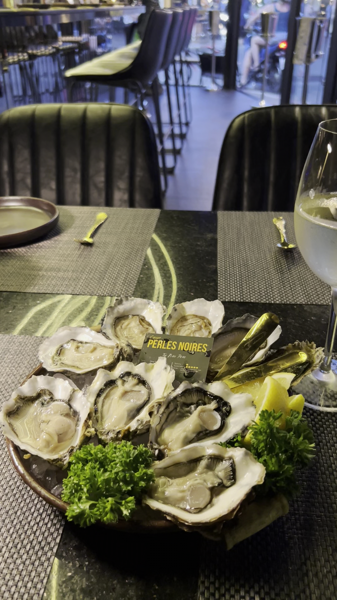จานหอยนางรมสดเสิร์ฟบนจานพร้อมผักชีฝรั่งและมะนาว ยี่ห้อ Perles Noires ในร้านอาหารหรูพร้อมแก้วไวน์ขาว