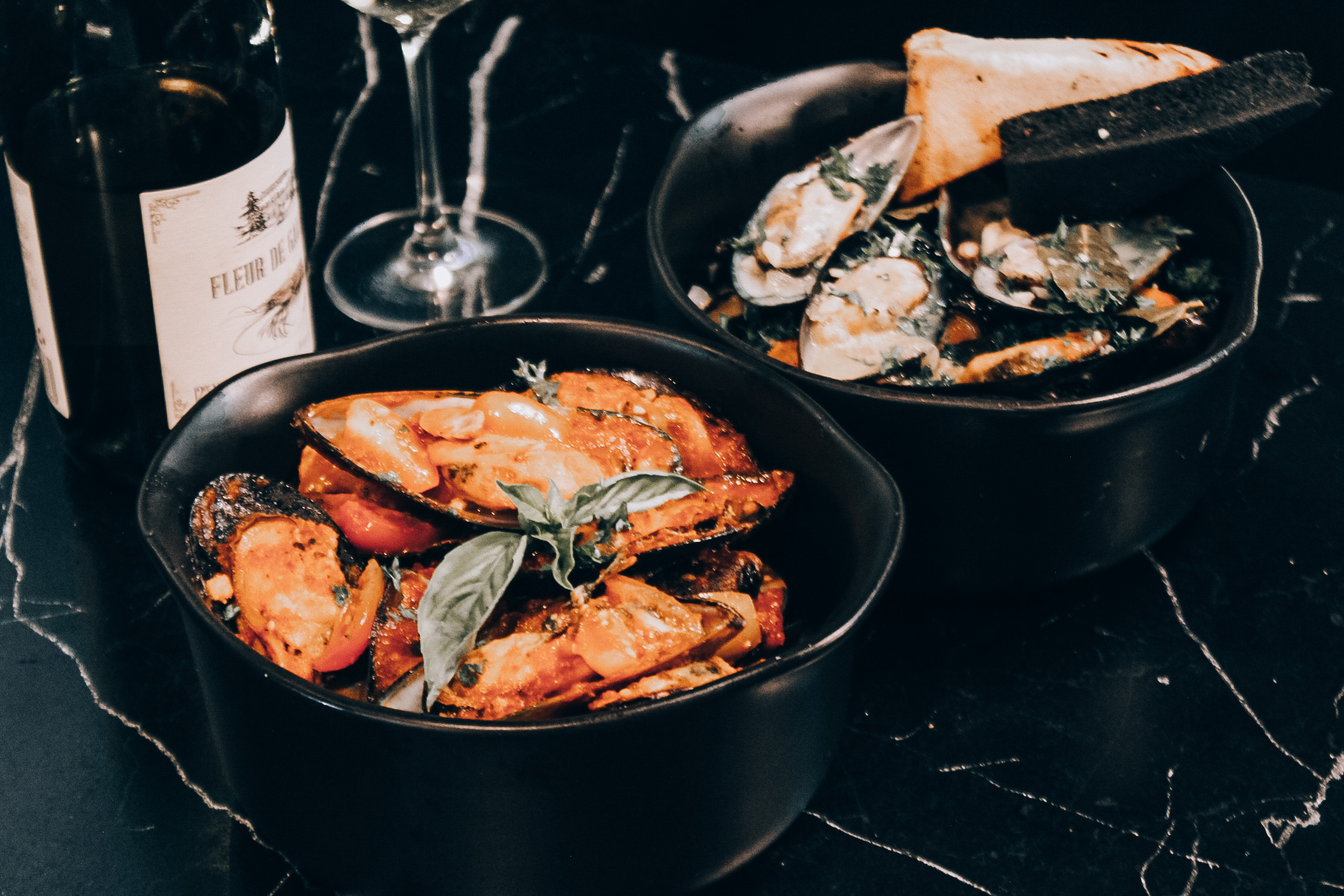 ชามหอยแมลงภู่ซอสมะเขือเทศสองชามพร้อมขนมปังและไวน์บนโต๊ะหินอ่อนสีดำ