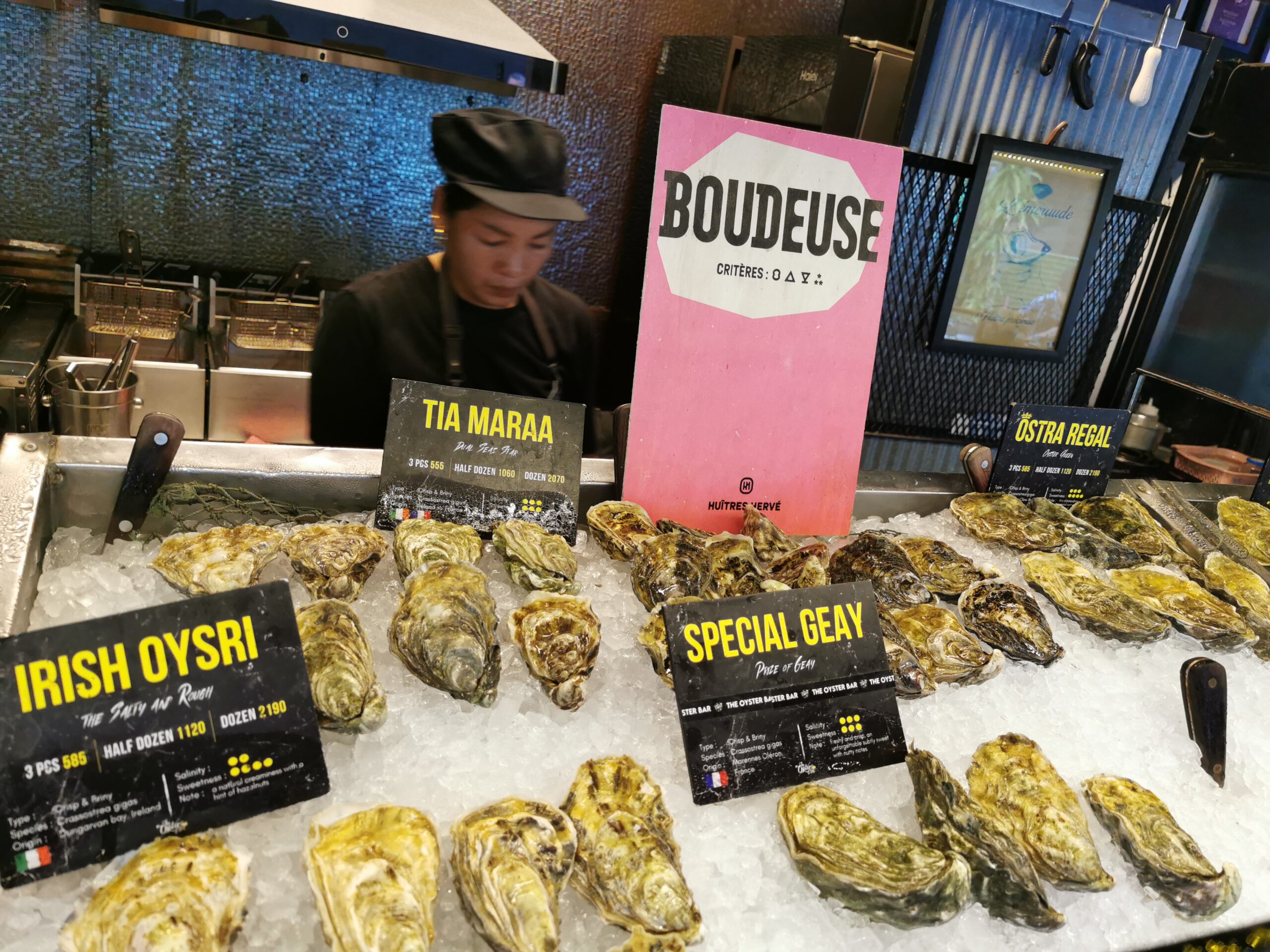 บาร์หอยนางรมสดพร้อมป้ายแสดงชนิดต่างๆ เช่น Irish Oysri, Tia Maraa, Special Geay และ Ostra Regal วางเรียงบนน้ำแข็ง พร้อมป้าย Boudeuse จาก Huîtres Hervé