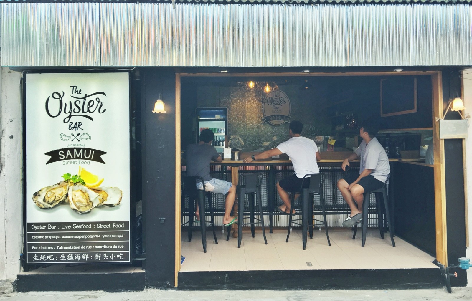 ร้าน The Oyster Bar สมุย บาร์อาหารทะเลสดและสตรีทฟู้ด มีลูกค้านั่งทานที่เคาน์เตอร์บาร์ ป้ายโฆษณาหอยนางรมสด