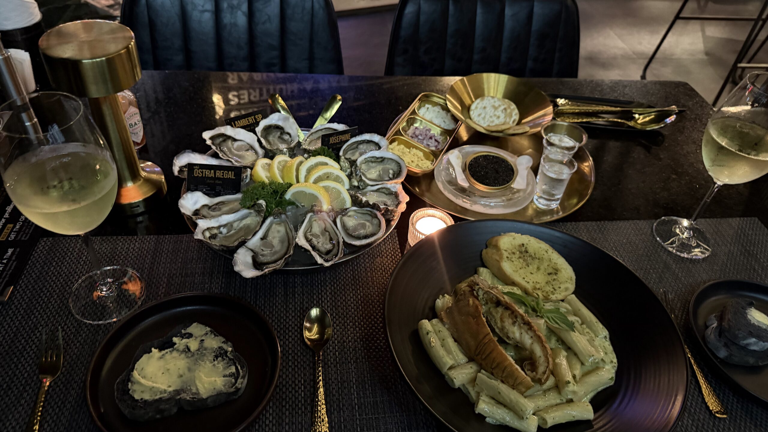 จานหอยนางรม Ostra Regal เสิร์ฟพร้อมมะนาว คาเวียร์ พาสต้าซอสครีมกุ้ง และไวน์ขาว บนโต๊ะอาหารสีดำตกแต่งด้วยภาชนะสีทอง