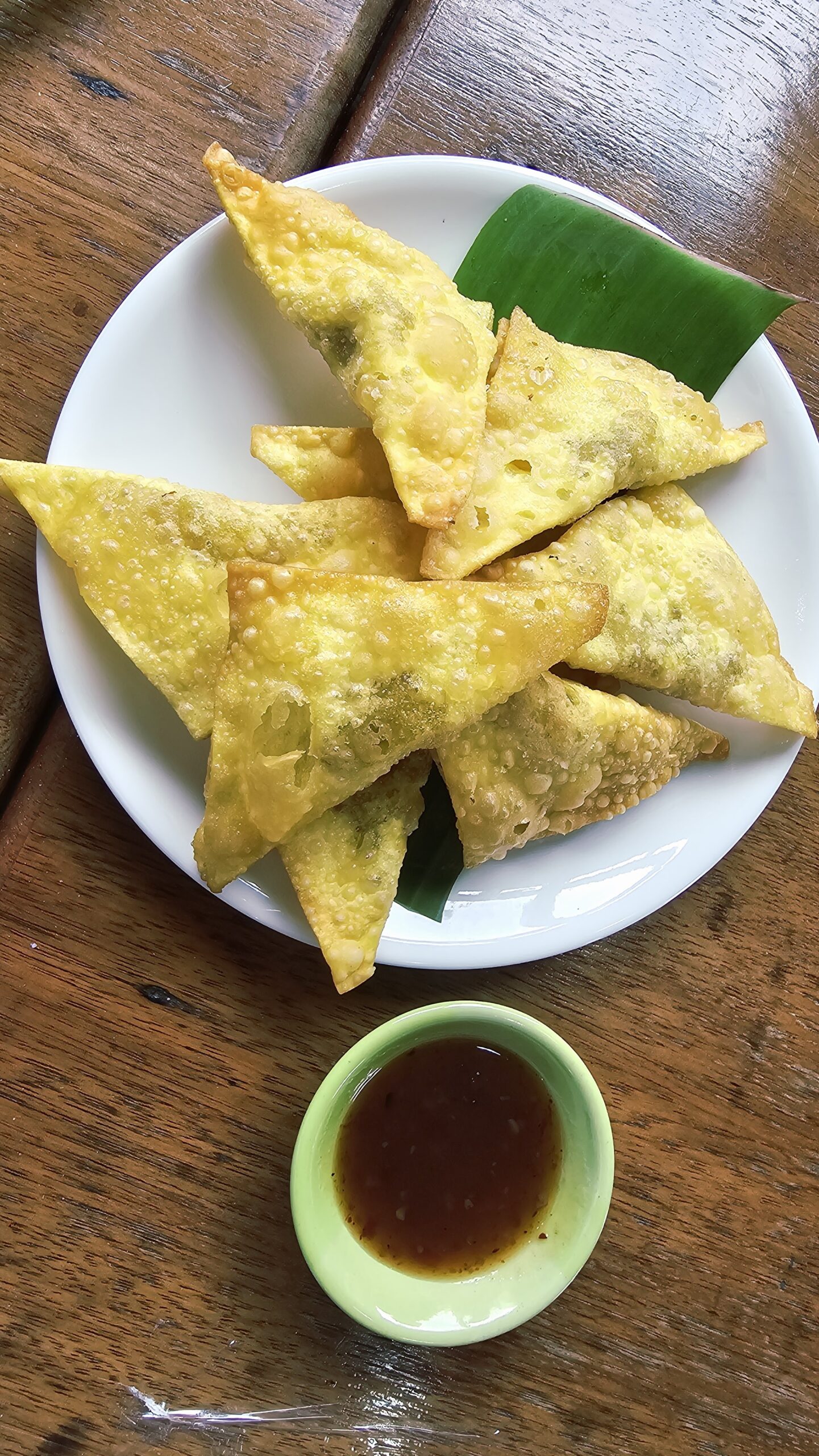เกี๊ยวทอดกรอบสีทองเสิร์ฟบนจานขาวรองใบตองพร้อมน้ำจิ้มเปรี้ยวหวานในถ้วยเขียวบนโต๊ะไม้