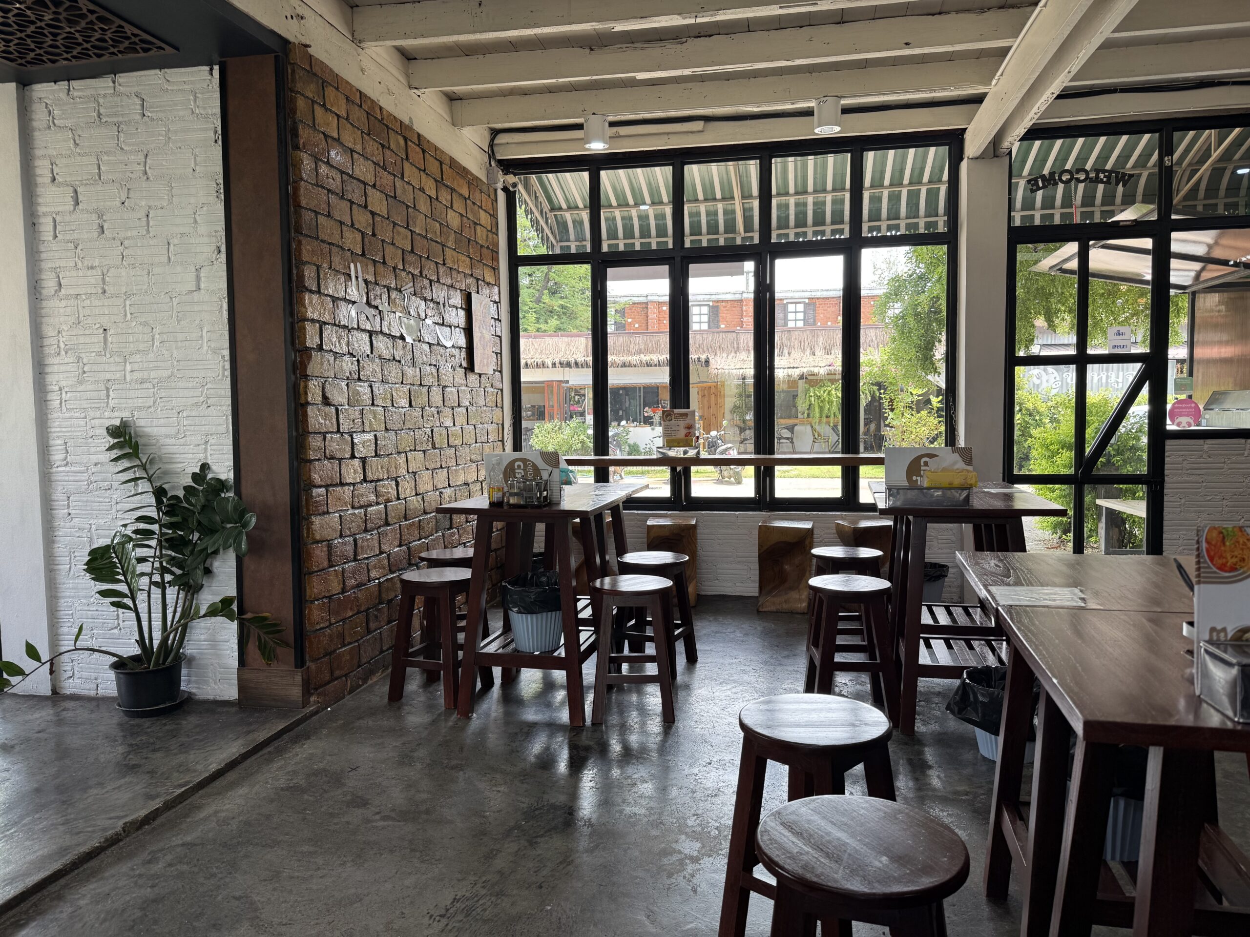 ภายในร้านอาหารสไตล์ลอฟท์ มีผนังอิฐแดง เก้าอี้สตูลไม้ โต๊ะไม้ หน้าต่างกระจกบานใหญ่ พื้นปูนขัดมัน และต้นไม้ประดับ