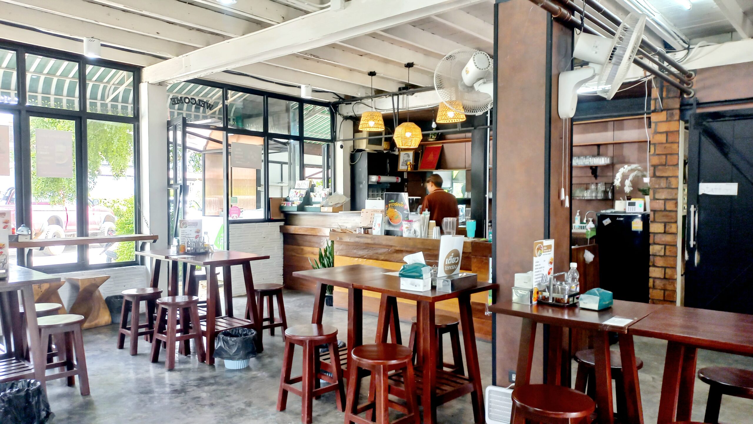 ภายในร้านอาหารสไตล์อินดัสเทรียล มีโต๊ะไม้สูงพร้อมเก้าอี้สตูลไม้สีเข้ม เคาน์เตอร์สั่งอาหาร โคมไฟหวาย พัดลมติดผนัง และหน้าต่างกระจกบานใหญ่