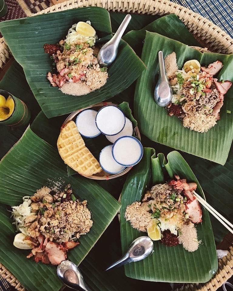อาหารไทยจัดเสิร์ฟบนใบตองสีเขียวบนถาดจักสาน มีเนื้อสับ ข้าวคั่ว มะนาว พริกป่น พร้อมขนมปังกรอบและเครื่องดื่มในถ้วยเซรามิก
