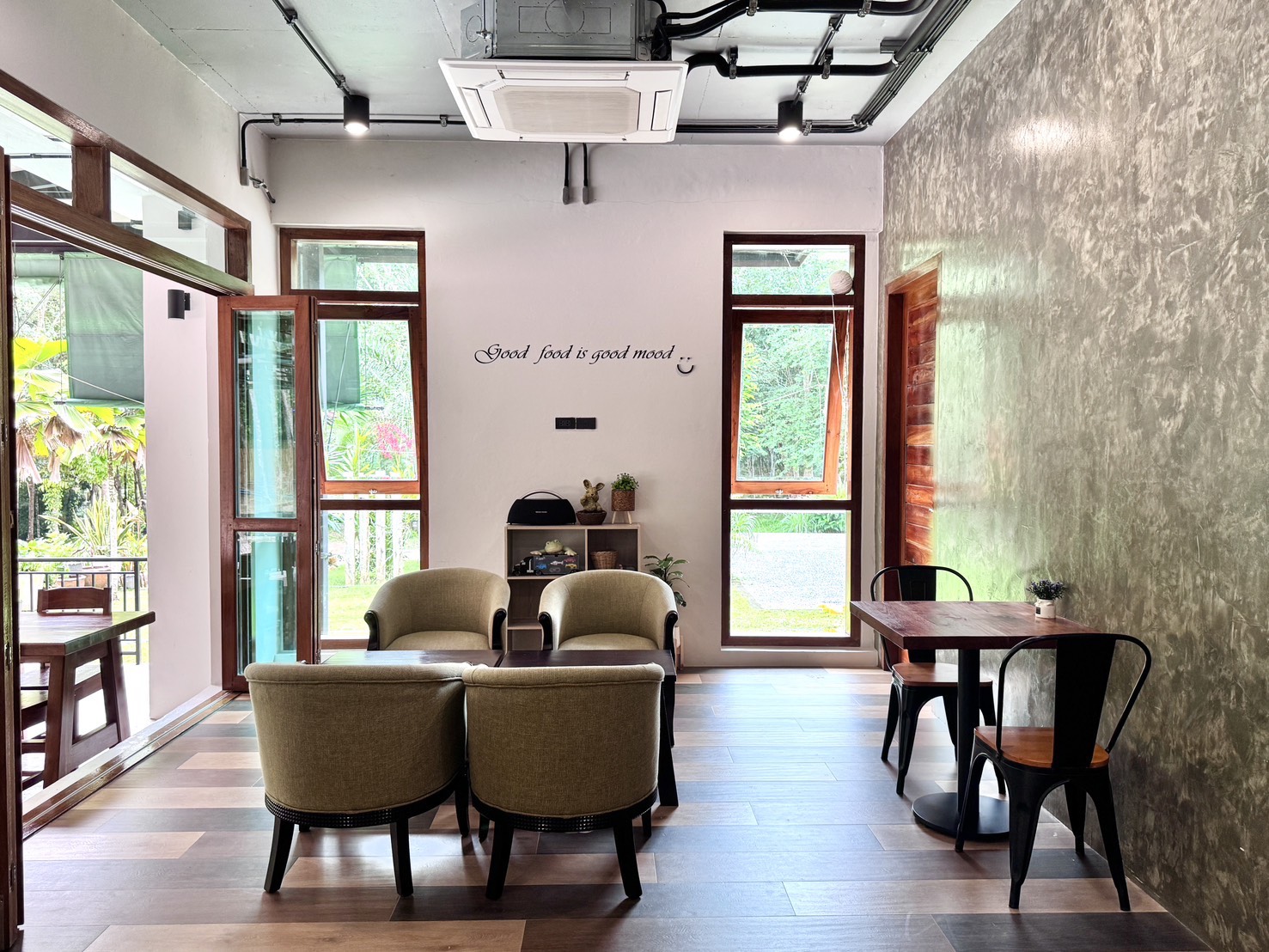 มุมนั่งเล่นภายในร้านอาหารสไตล์คาเฟ่ มีเก้าอี้โซฟาสีเขียวมะกอก โต๊ะไม้ เก้าอี้เหล็กสีดำ ผนังปูนเปลือย หน้าต่างกรอบไม้ และข้อความบนผนังว่า Good food is good mood