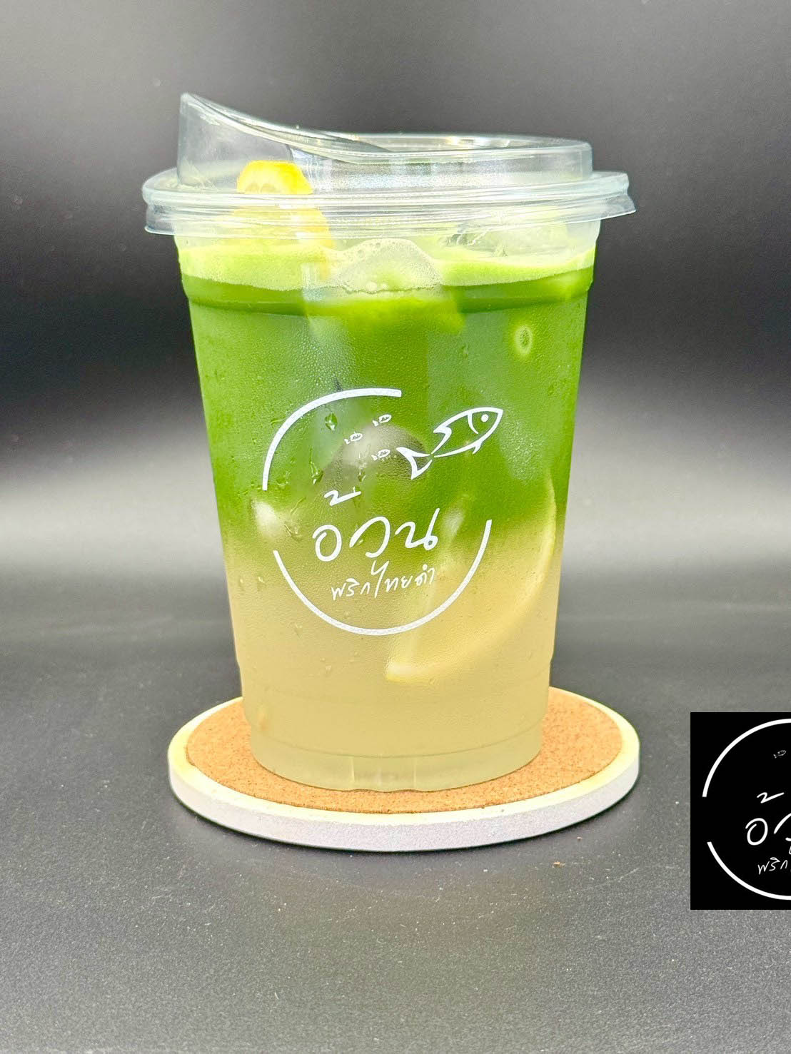 เครื่องดื่มชาเขียวมัทฉะเย็นในแก้วพลาสติกพร้อมโลโก้ร้านอวนพริกไทยดำ วางบนจานรองไม้ก๊อก
