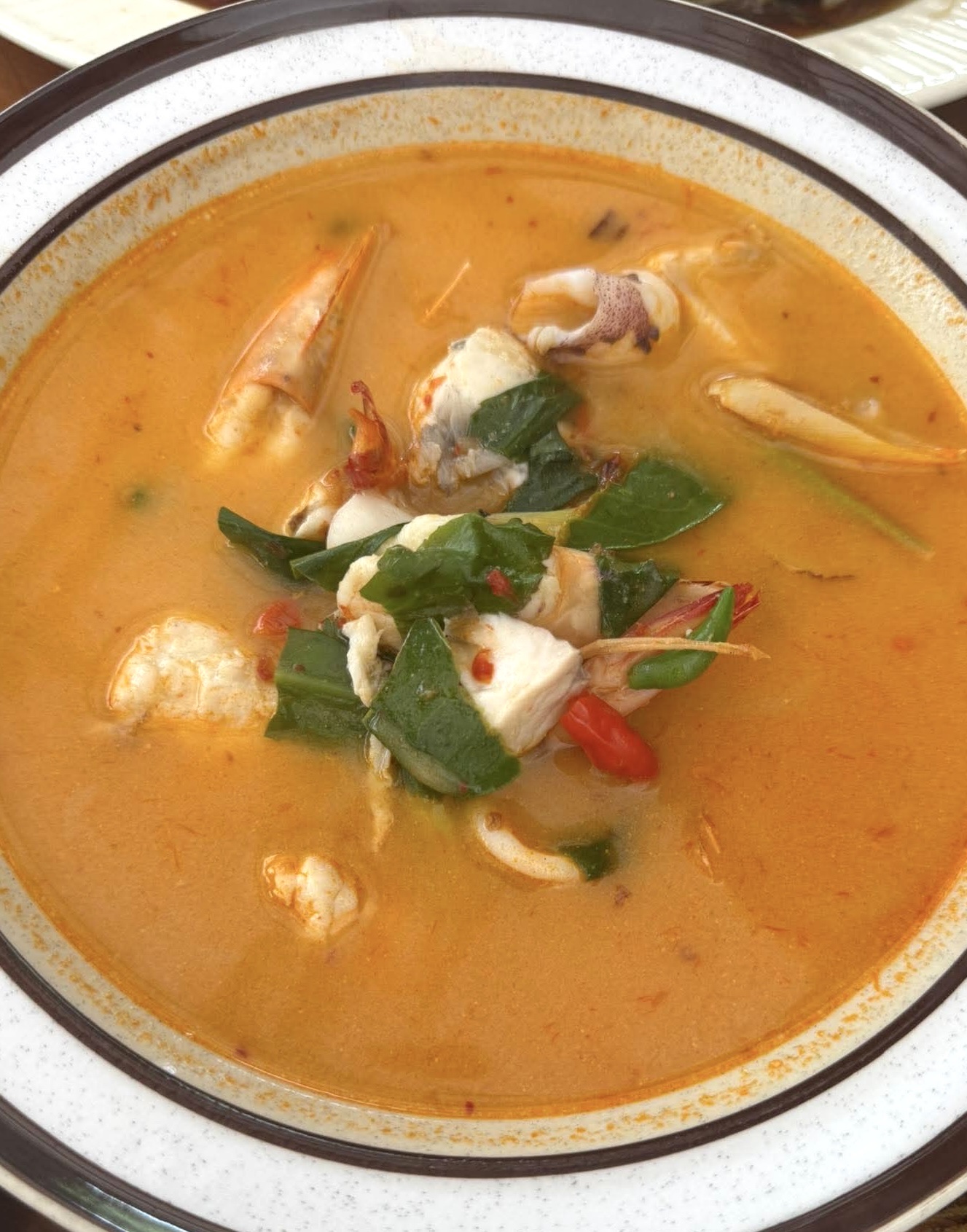 ต้มยำกุ้งน้ำข้นใส่ปูและอาหารทะเล เสิร์ฟในชามขอบดำตกแต่งด้วยใบโหระพาและพริก