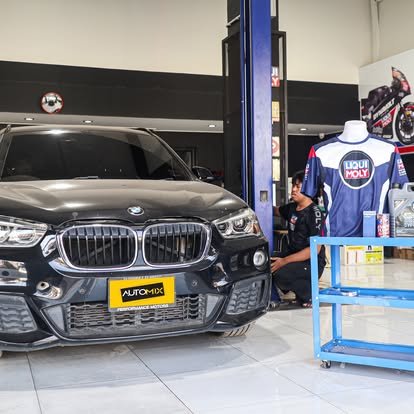 รถ BMW สีเทาเข้มจอดอยู่ในศูนย์บริการรถยนต์ Auto Mix พร้อมช่างกำลังทำงานและชุดยูนิฟอร์ม Liqui Moly แขวนอยู่