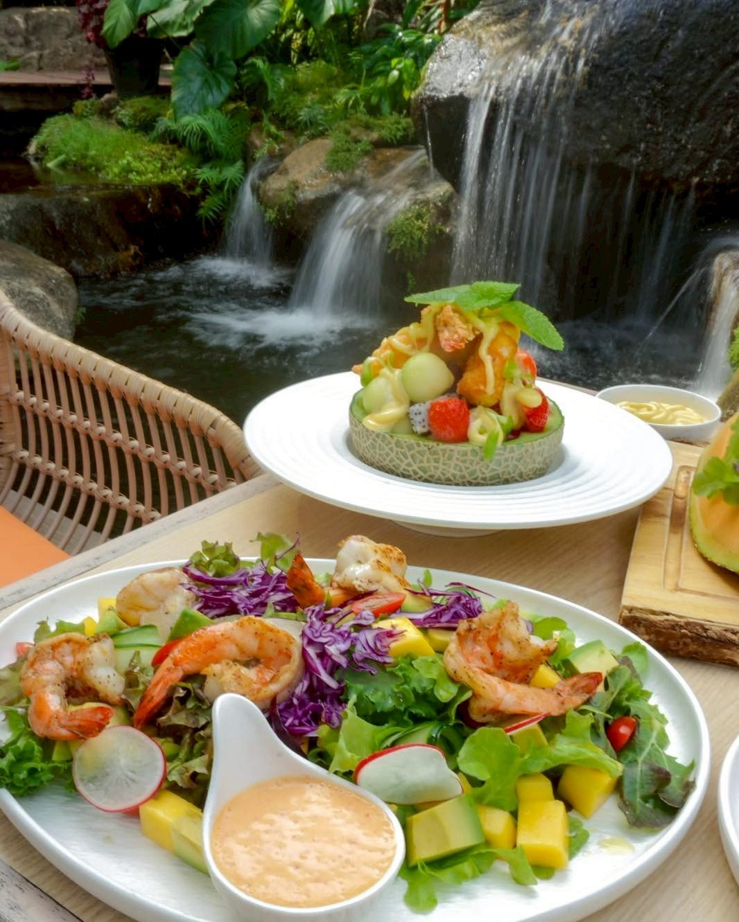 สลัดกุ้งสดพร้อมผักและมะม่วง เสิร์ฟคู่กับผลไม้รวมในเมลอนแกะสลัก บนโต๊ะอาหารริมน้ำตกในร้านอาหารบรรยากาศธรรมชาติ