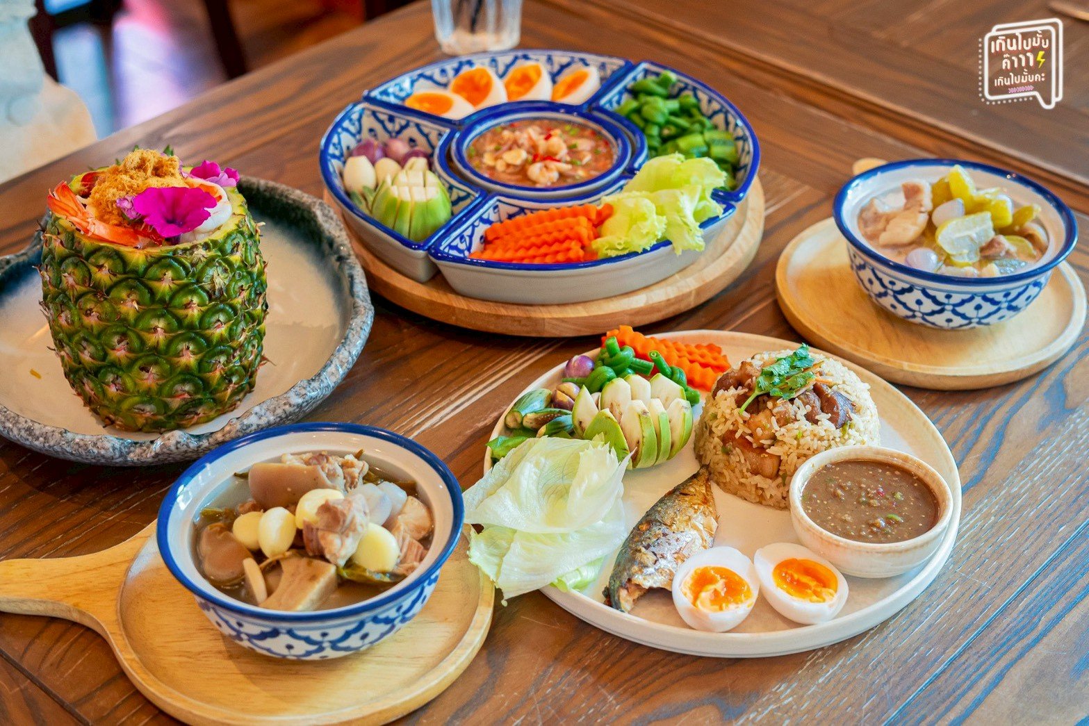 สำรับอาหารไทยโบราณบนโต๊ะไม้ มีข้าวผัดปลาทู น้ำพริกกะปิผักสด แกงจืด ข้าวอบสับปะรด และขนมหวาน เสิร์ฟในถ้วยชามลายครามสีน้ำเงินขาว