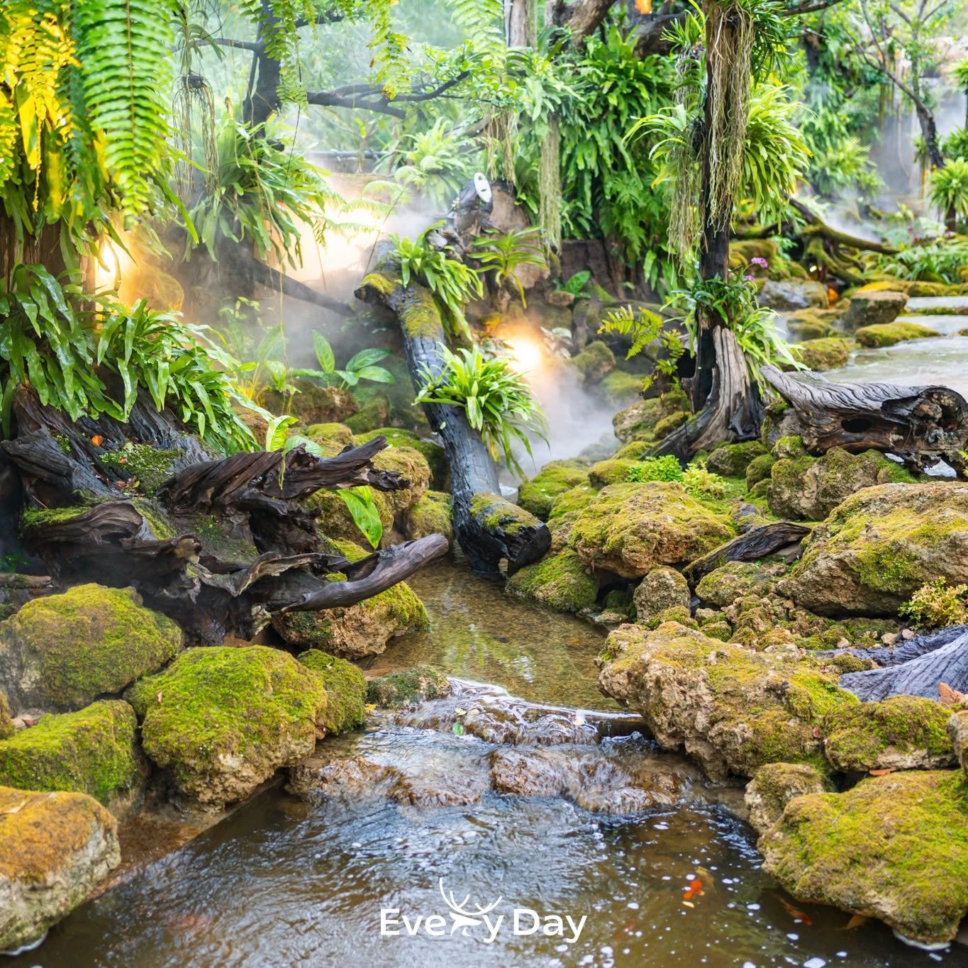 สวนเขตร้อนในร้านอาหารที่มีลำธารน้ำไหลผ่านหินปกคลุมด้วยมอส ล้อมรอบด้วยเฟิร์นและไม้เขียวชอุ่ม พร้อมหมอกควันและแสงไฟสวยงาม