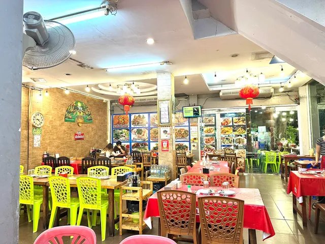 ภายในร้านอาหารไทยที่มีเก้าอี้สีสันสดใส โคมไฟจีนสีแดง เมนูอาหารติดผนัง และพัดลมติดเพดาน บรรยากาศแบบร้านอาหารริมถนน
